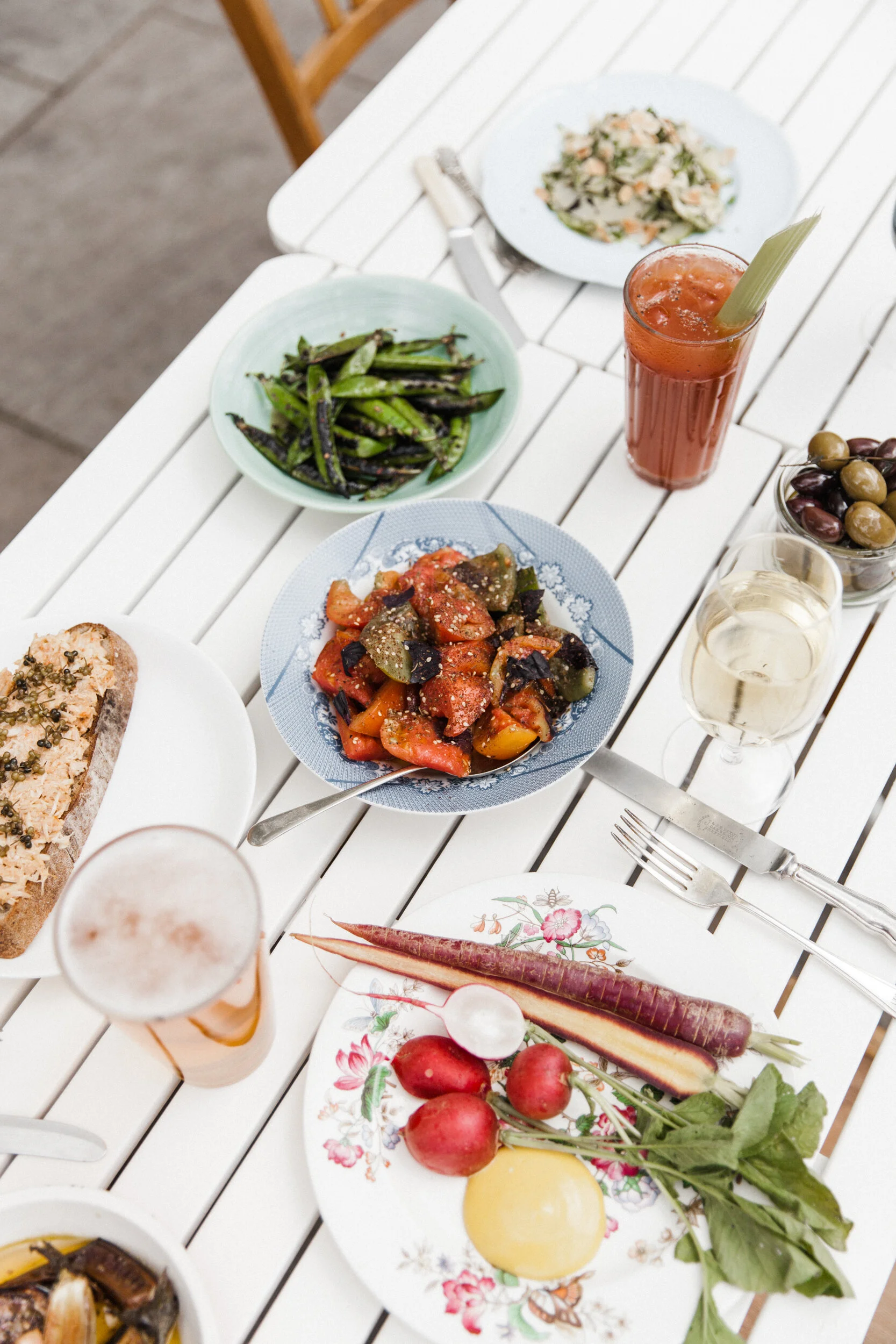 A white table with various dishes, including a bowl of grilled green beans, a plate of ratatouille, a bowl of mixed olives, a sliced baguette, radishes with greens, and a glass of pink drink with a straw.