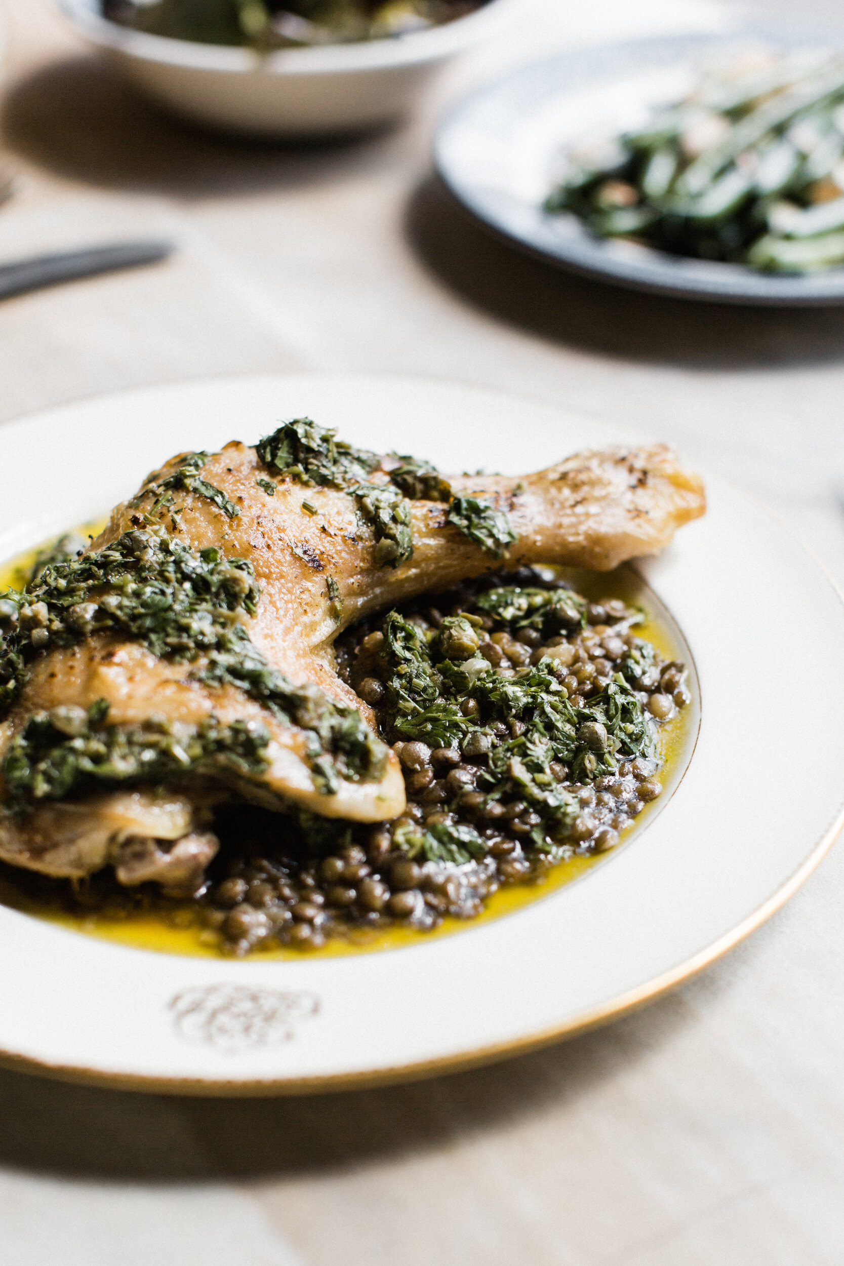 A plate with cooked chicken topped with herbs, served with a bed of cooked lentils with herbs in a sauce, on a white decorative dinner plate.