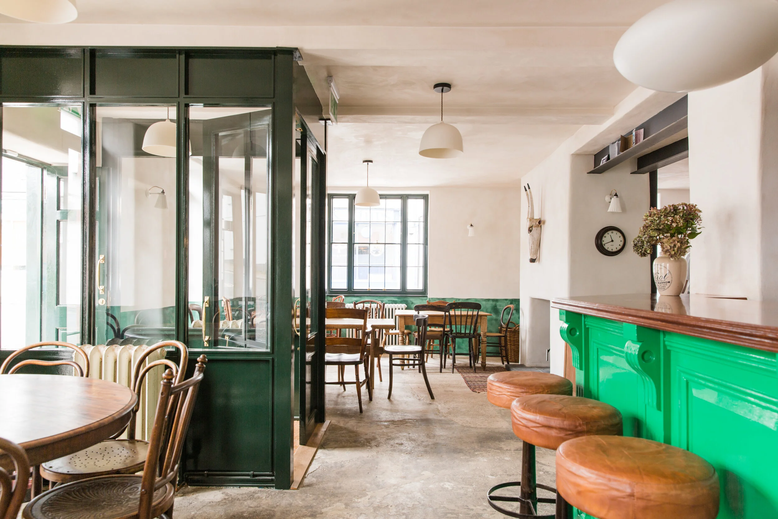 Interior of a cafe with wooden tables and chairs, a distinctive green bar counter, a white vase with flowers, multiple windows letting in natural light, and modern hanging pendant lights.