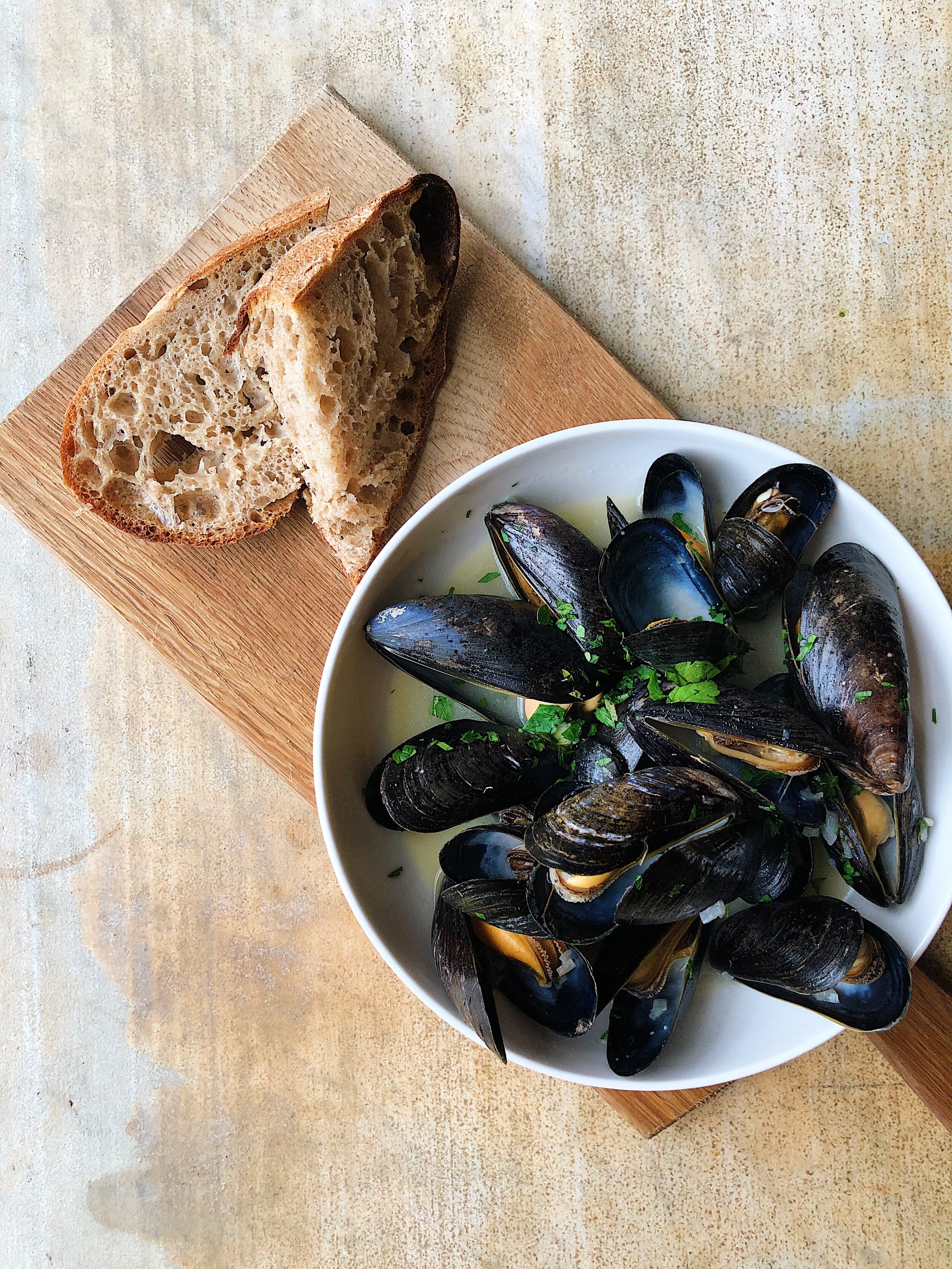 A bowl of cooked mussels garnished with chopped parsley placed next to two slices of rustic bread on a wooden cutting board.