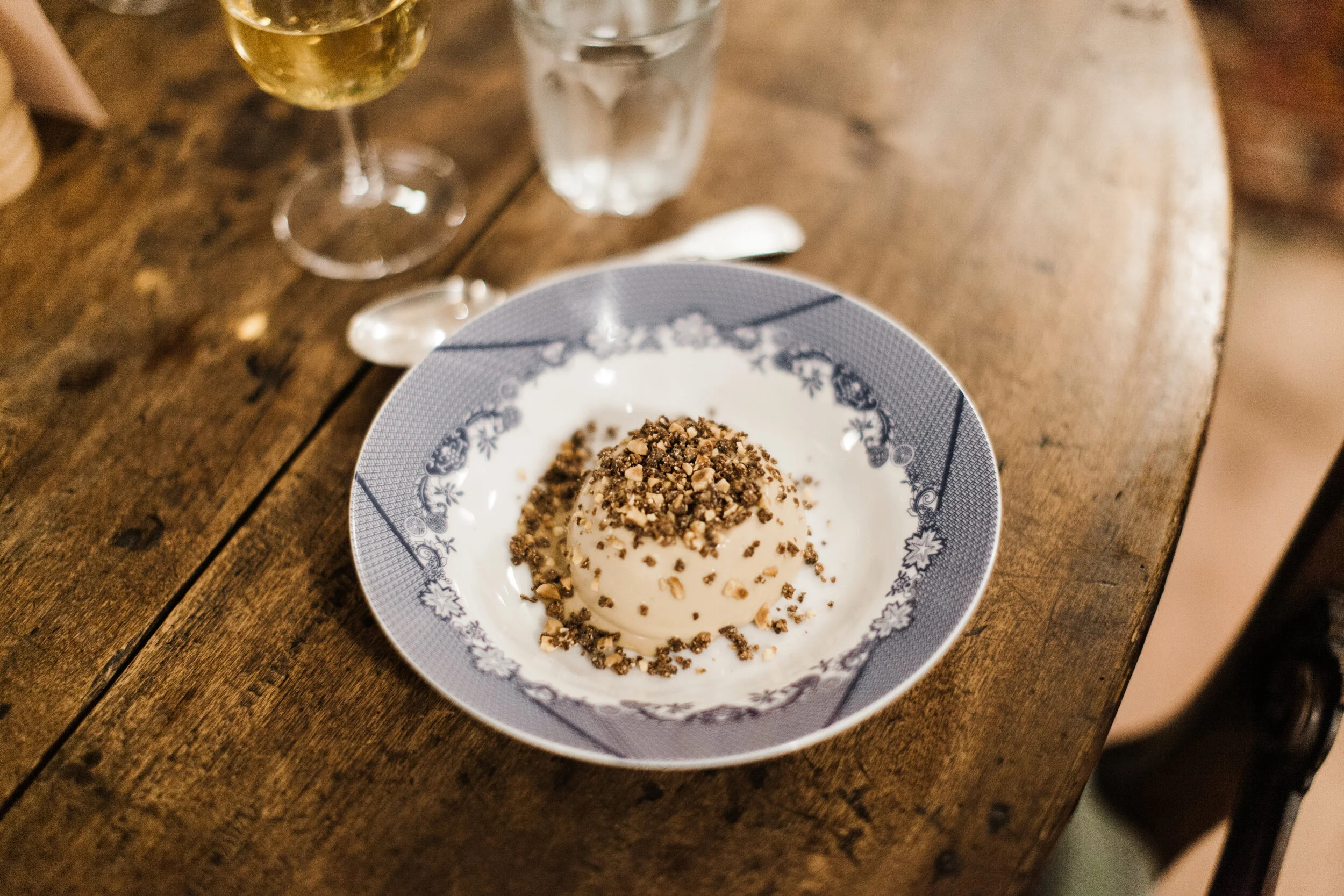 A bowl of ice cream topped with chopped nuts, placed on a wooden table. In the background, there are two glasses, one with a yellow beverage and the other with water, along with a spoon.