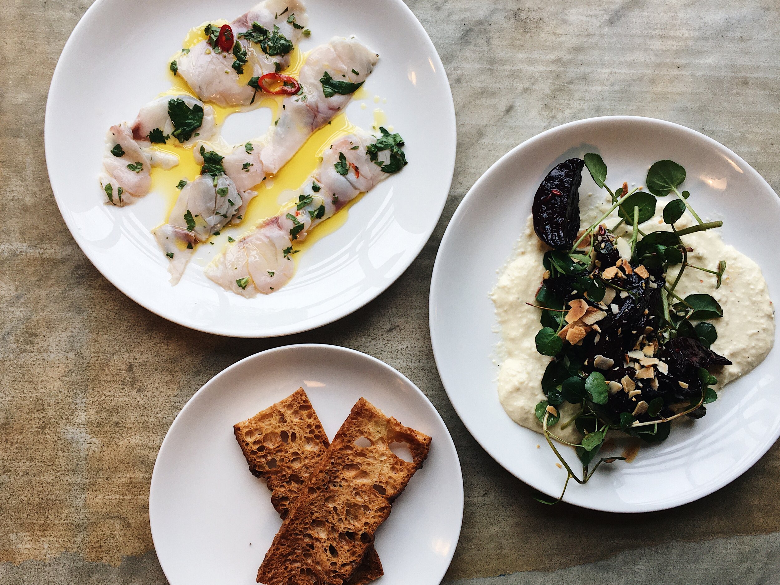 Three white plates with different appetizers on a wooden table. The top plate has raw fish slices with herbs, chili, and olive oil. The middle plate has a dip with dark chutney, microgreens, and chopped nuts. The bottom plate has toasted bread slices