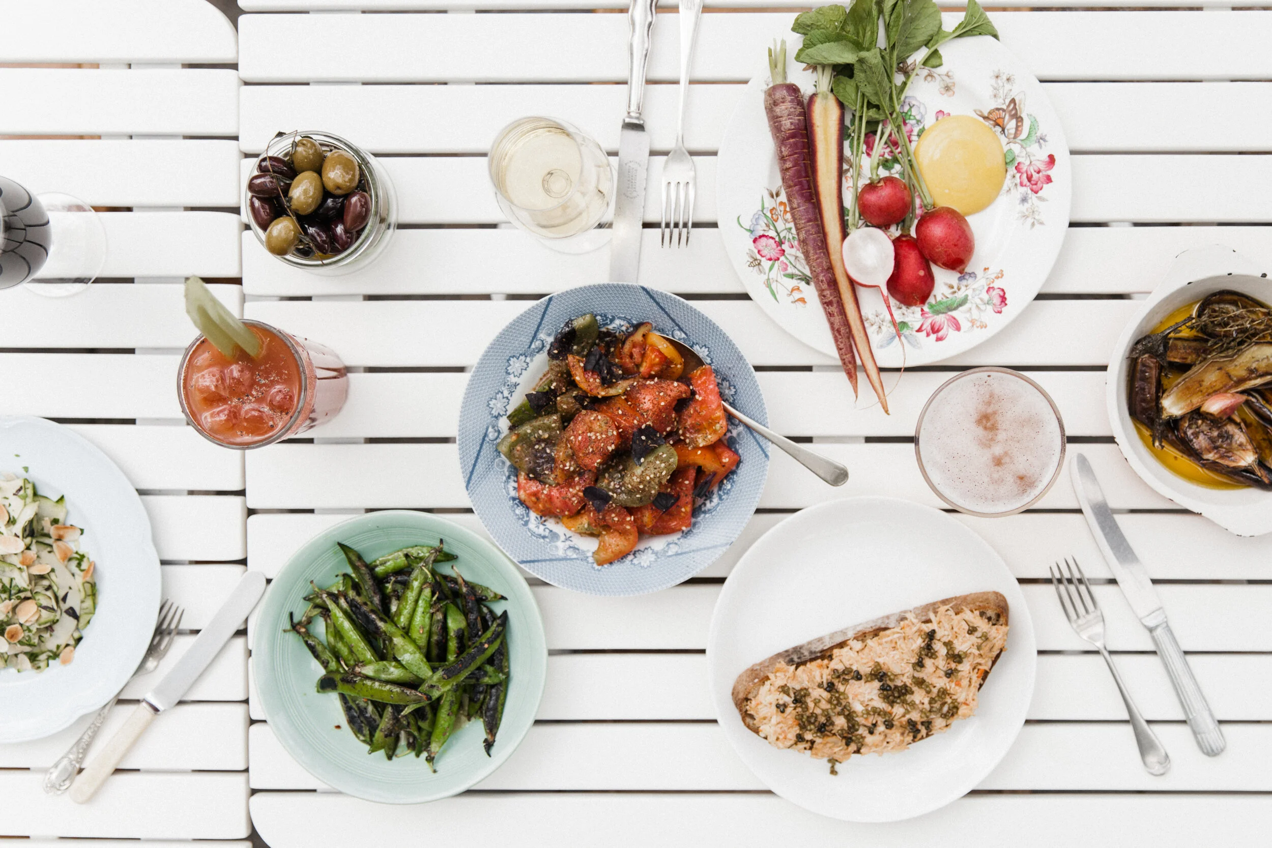 Top-down view of a table with various dishes including a salad, grilled vegetables, a slice of toasted bread with toppings, a bowl of olives, a glass of white wine, and a couple of glasses of pink and white drinks on a white wooden table.