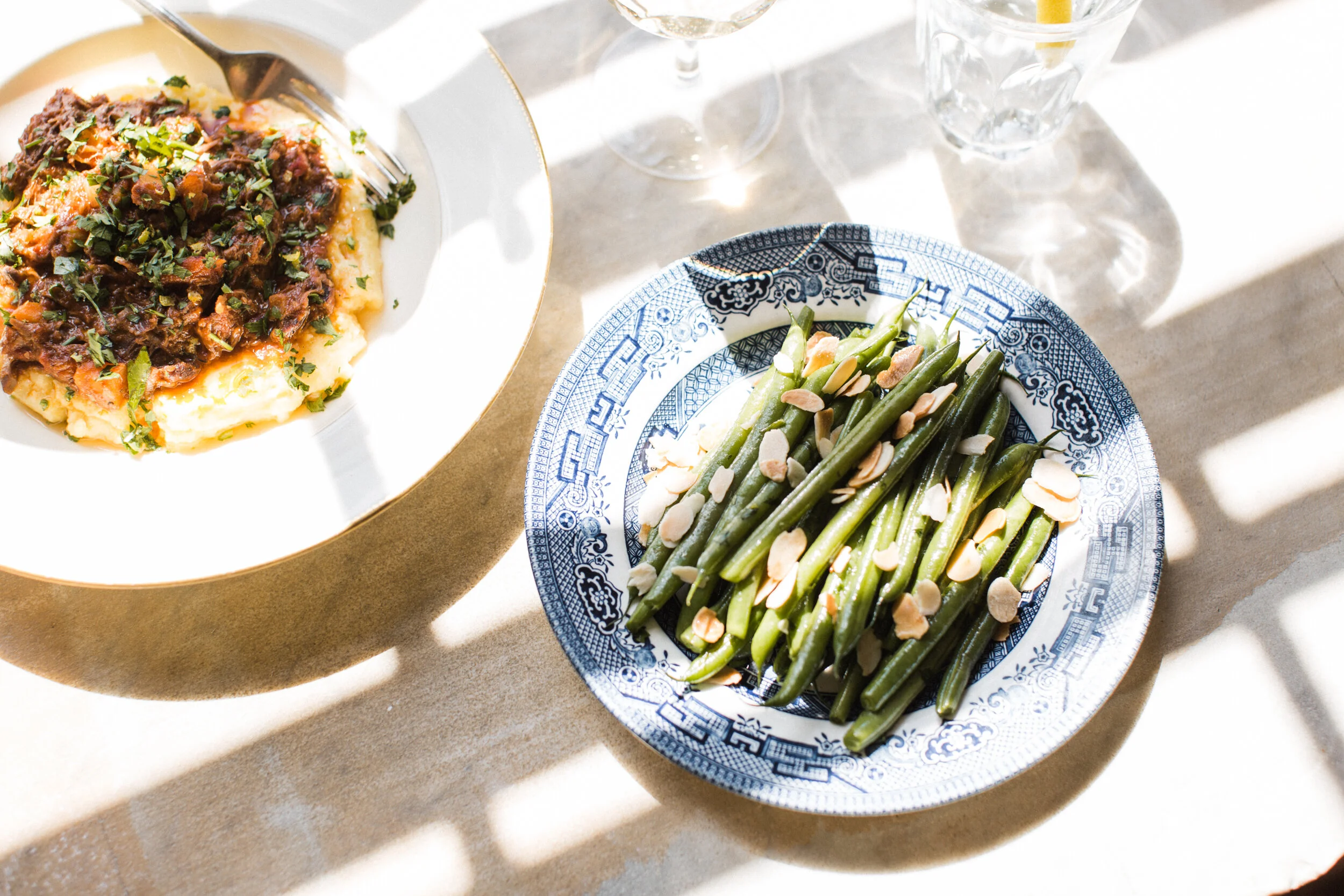 A plate of steak topped with herbs, served with mashed potatoes, and a side of green beans with sliced almonds. There are also two glasses of white wine on the table.