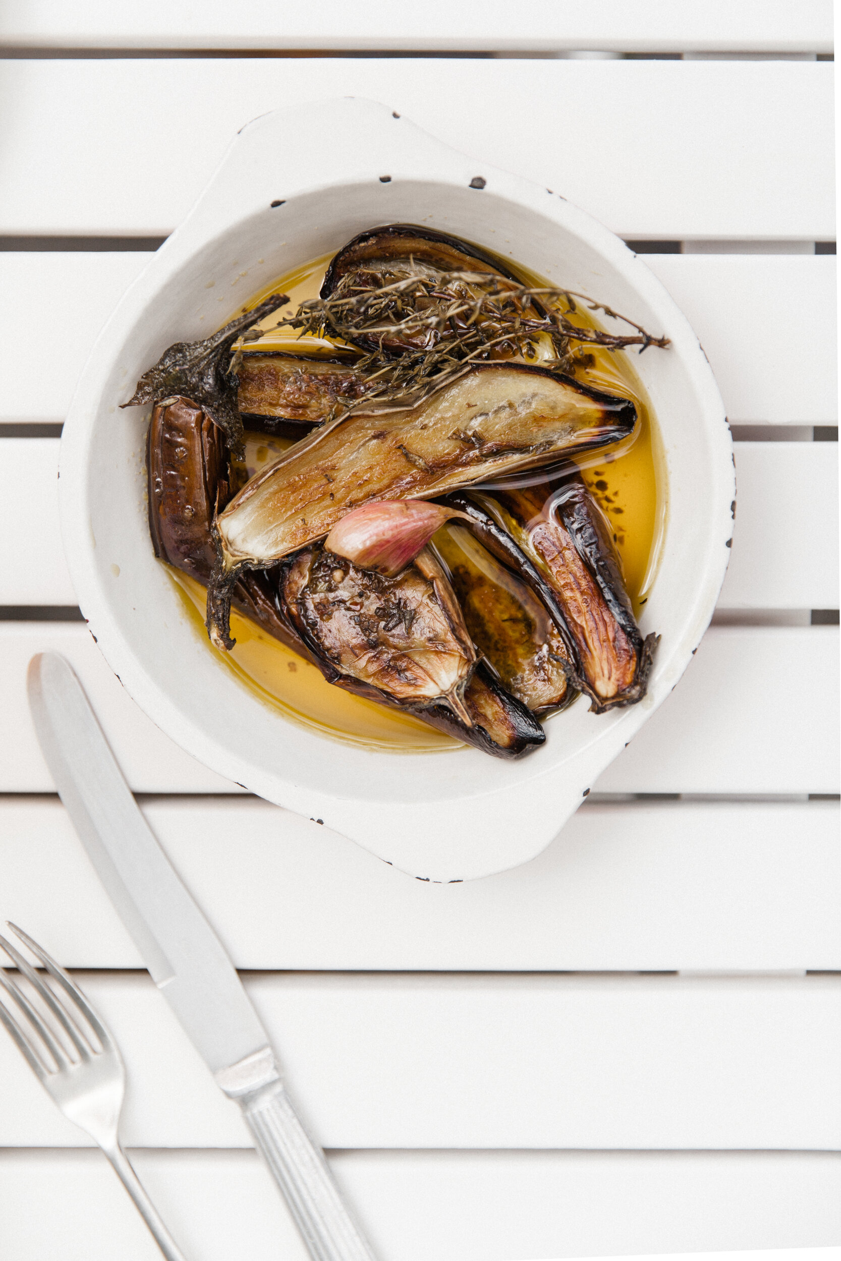 A white bowl containing roasted eggplants in oil, garnished with herbs and garlic, placed on a white slatted surface.