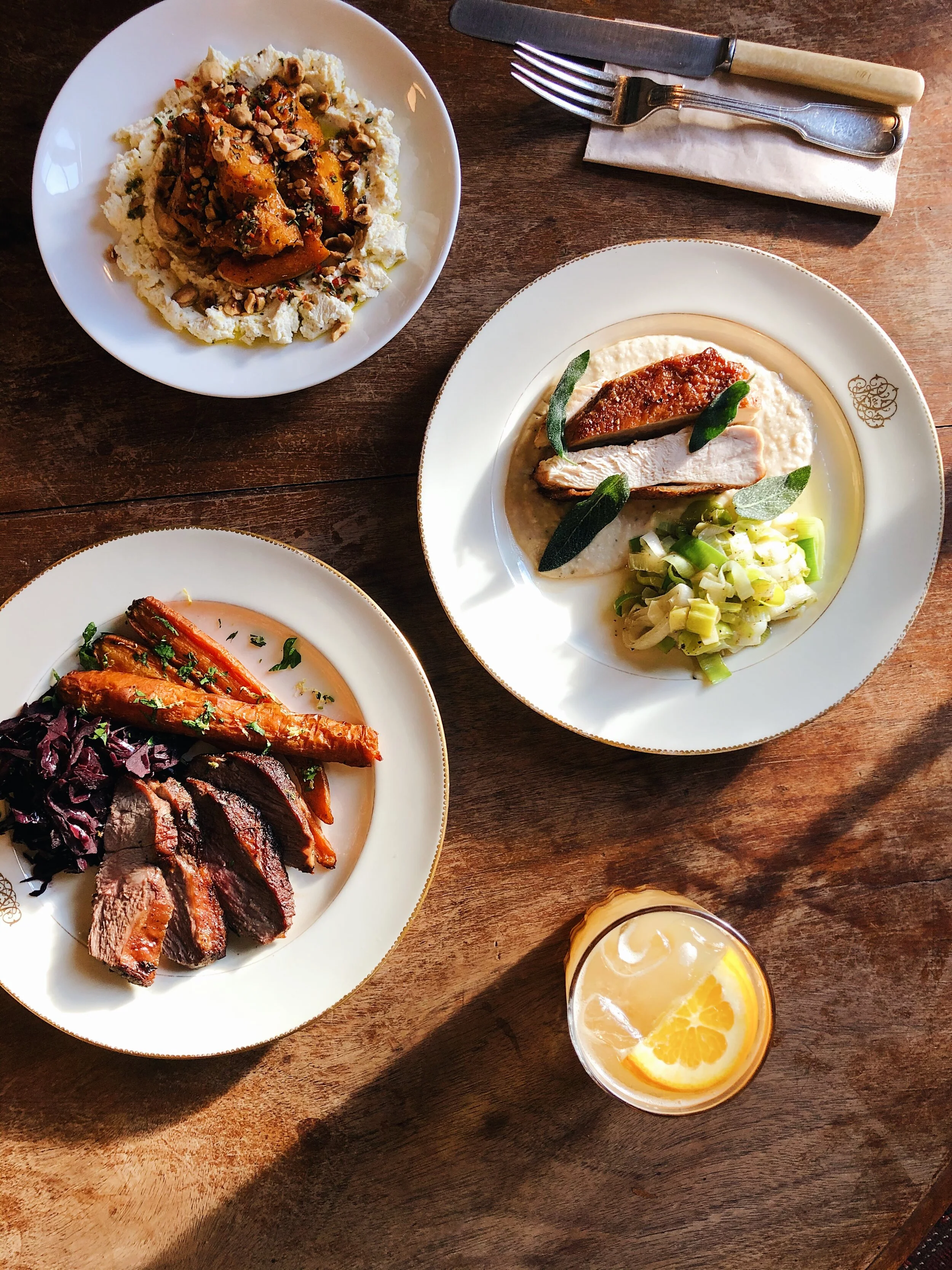 A table with plates of various foods including sliced meat, roasted carrots, greens, and a dish topped with nuts, along with a glass of lemon-infused beverage.