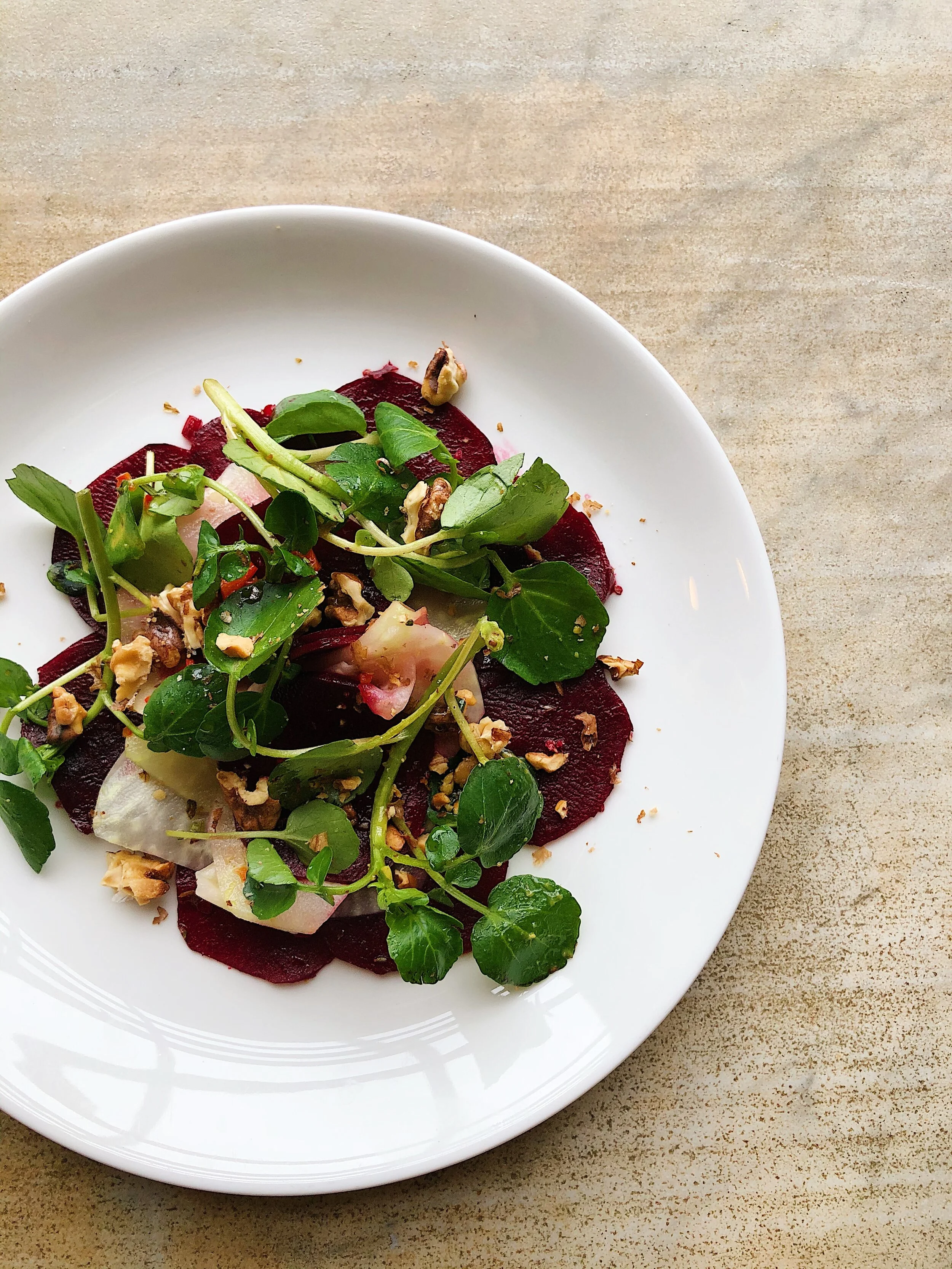Salad with greens, beets, walnuts, and sliced radishes on a white plate on a beige surface.