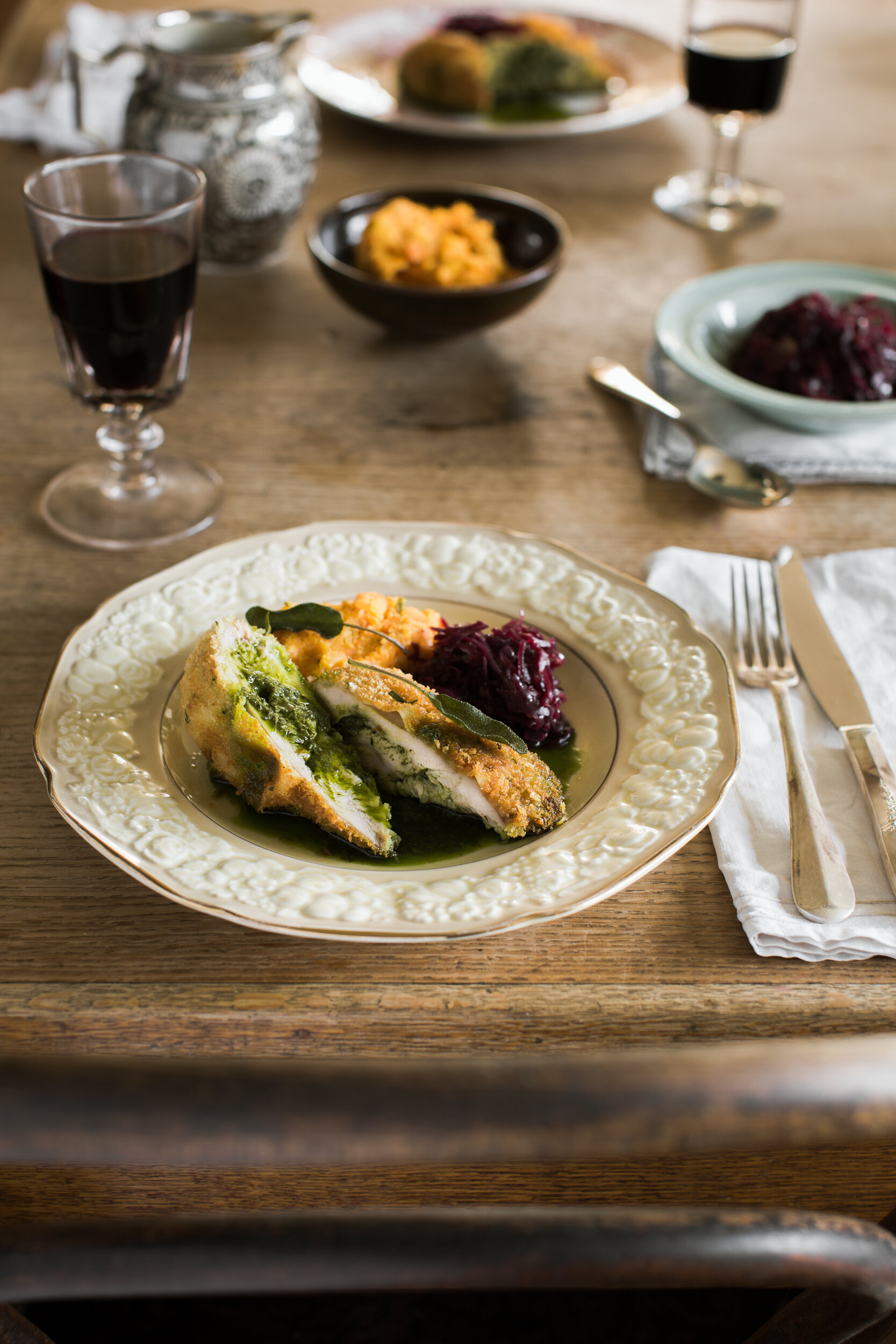 A plate with stuffed chicken breast, mashed potatoes, and cranberry sauce on a wooden table. In the background, there are glasses of red wine, bowls of side dishes, and a table set with silverware and cloth napkins.