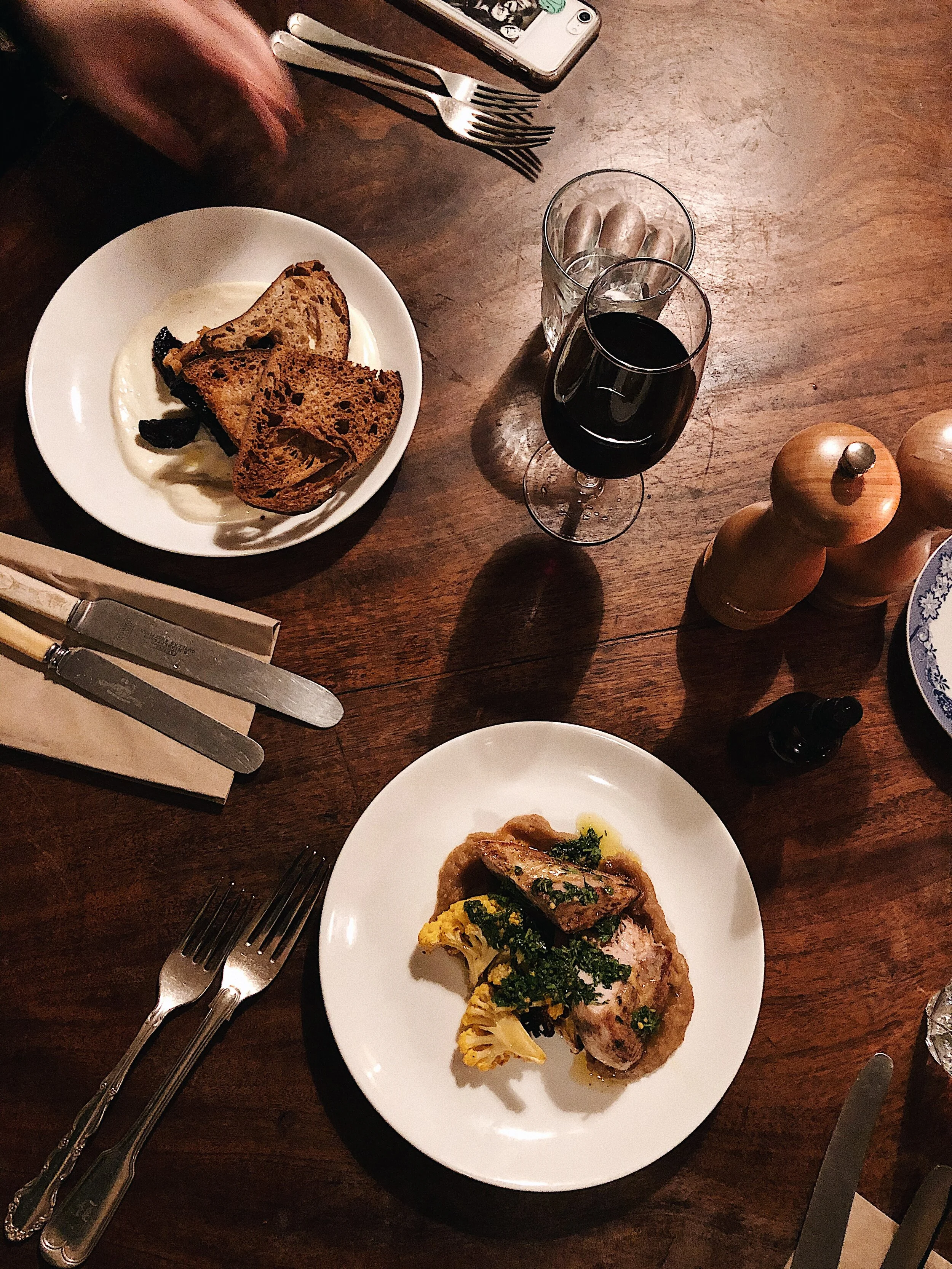 A wooden table with a plate of roasted chicken garnished with herbs and vegetables, a glass of red wine, and a glass of water. Silverware and a salt and pepper shaker are also on the table.