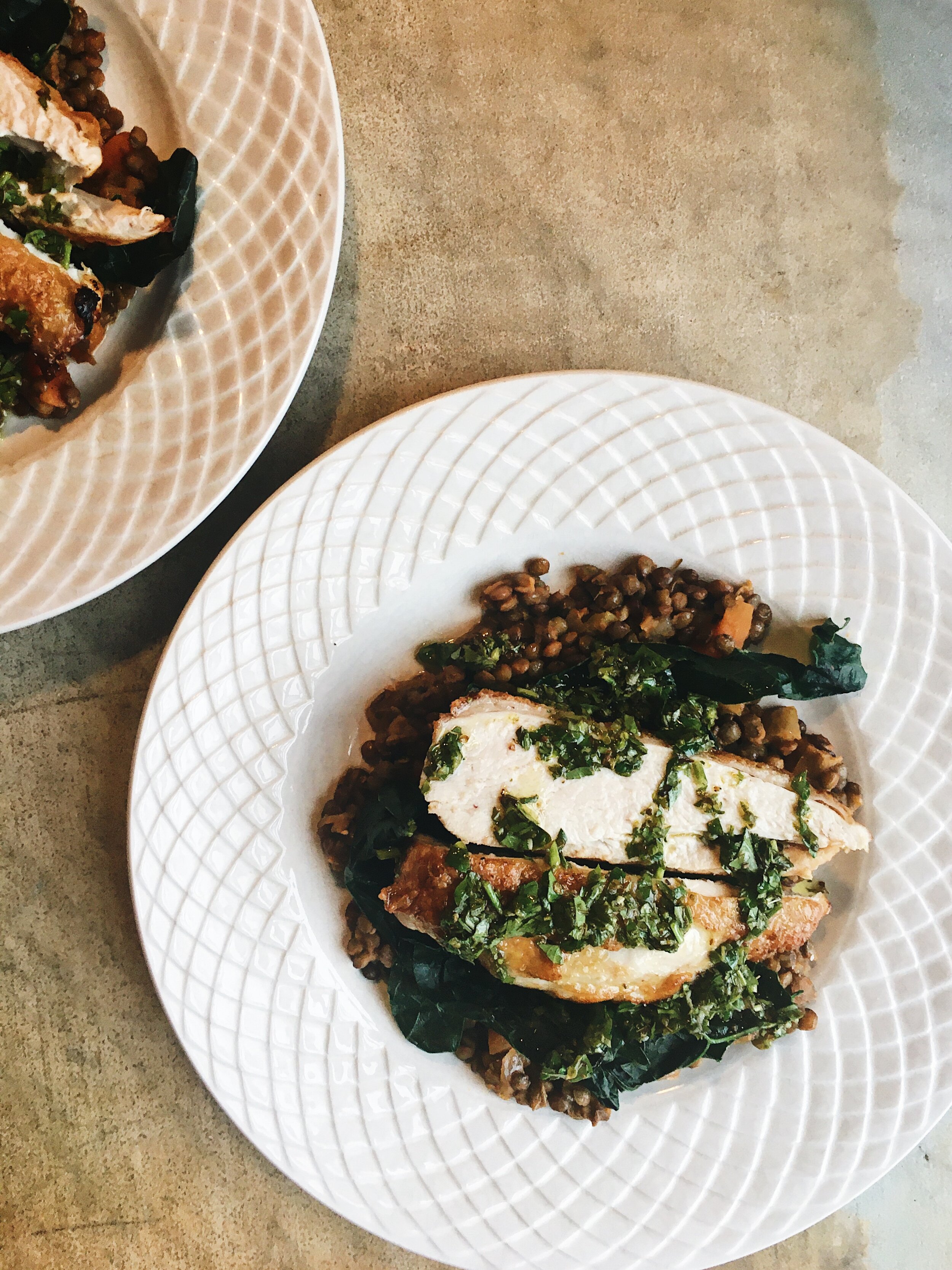 A white plate with a textured pattern holding a cooked fish fillet topped with chopped herbs, served over a bed of dark leafy greens and lentils, with some additional lentils on a second white plate nearby.