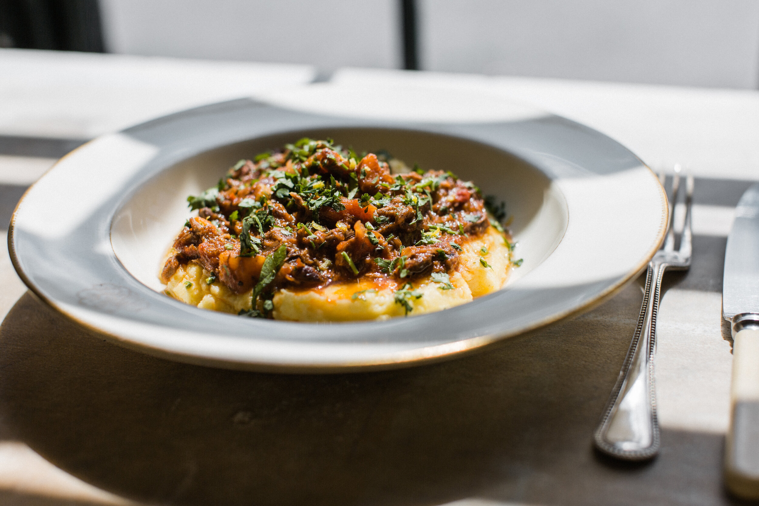 A plate of mashed potatoes topped with beef stew and garnished with chopped herbs, placed on a dark wooden table with a fork and knife beside it.