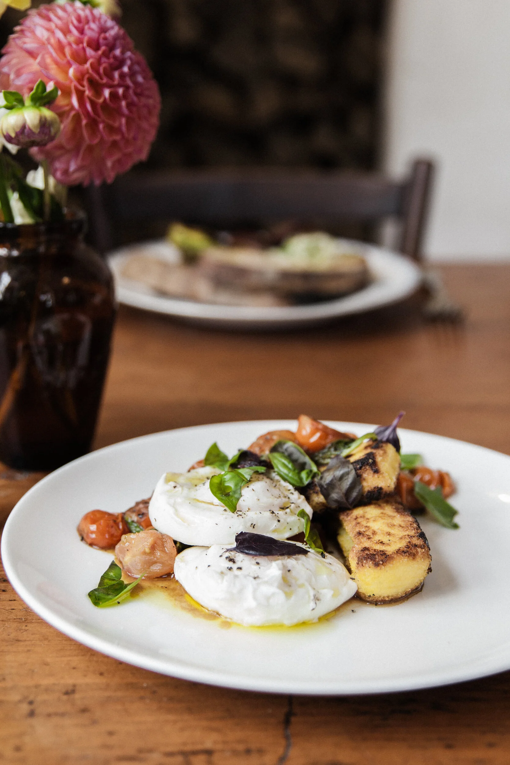 A white plate of caprese salad with fresh mozzarella, tomatoes, basil, grilled bread, and greens, with a blurred dining setting in the background.