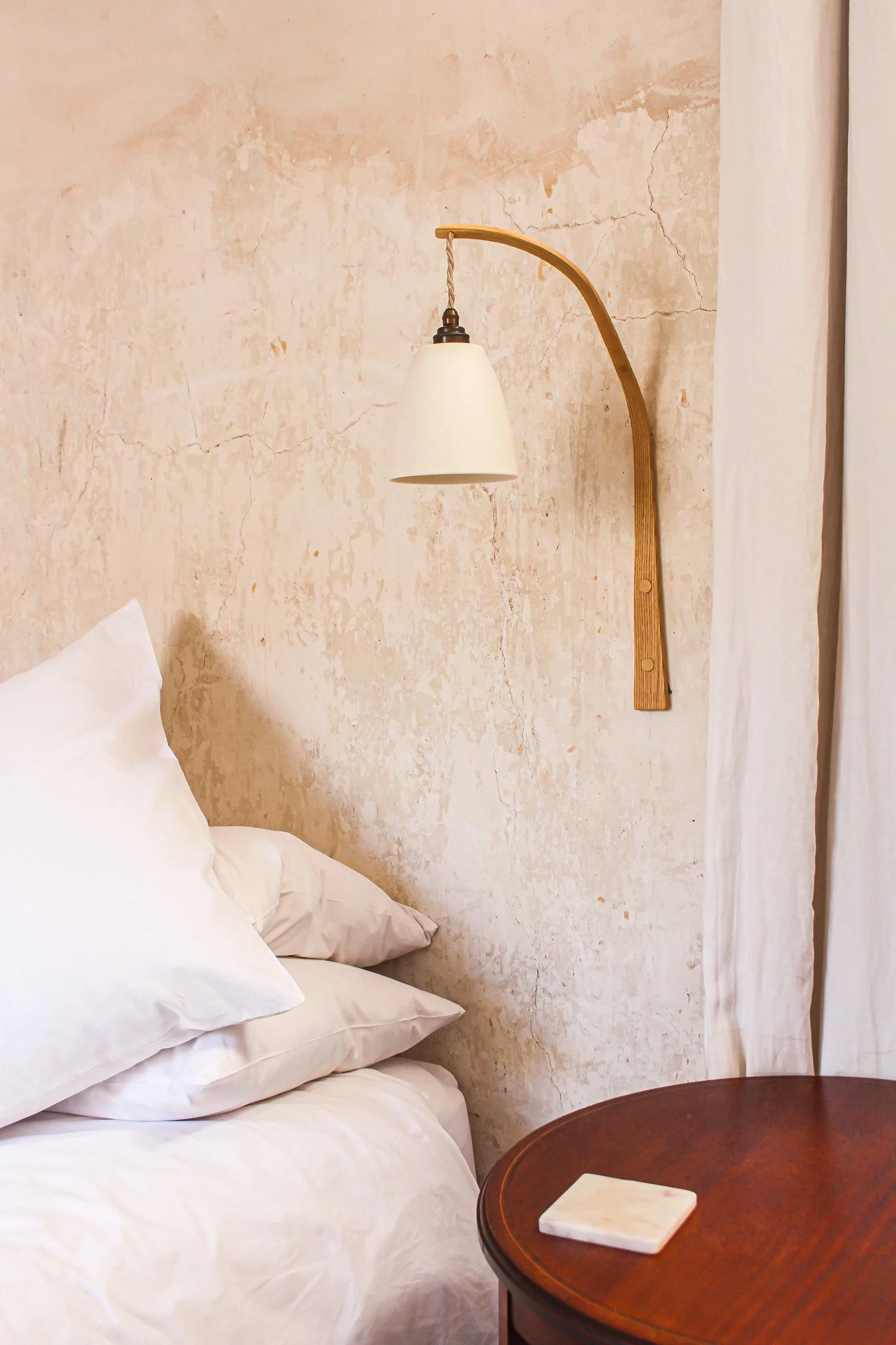 Part of a bedroom with a bed, white pillows, a bedside wall lamp with a curved wooden arm, a textured plaster wall, and a round wooden table with a marble-patterned notepad.