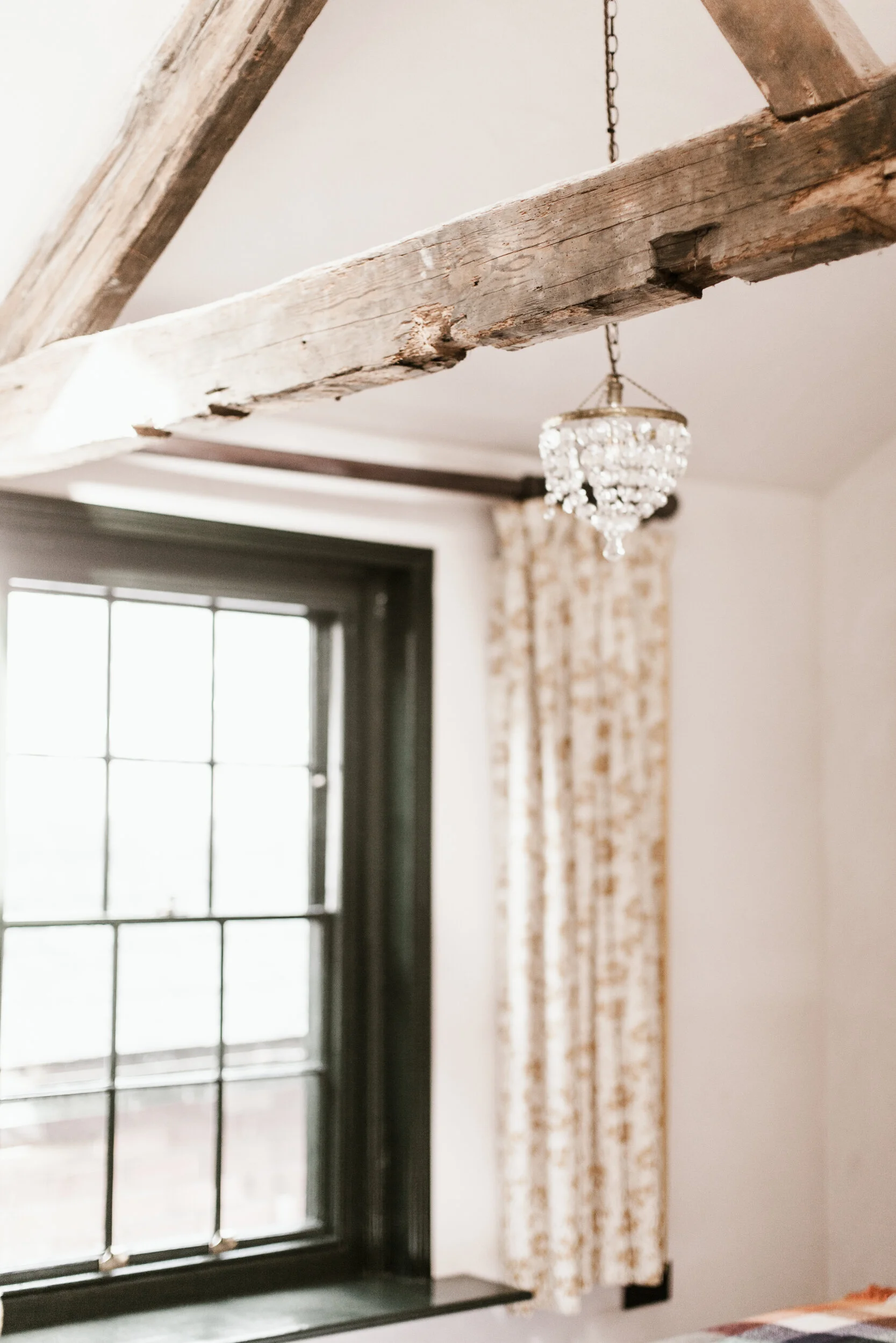 Interior view of a room with a large window with black framing, beige patterned curtains, a wooden beam on the ceiling, and a chandelier hanging from a chain.