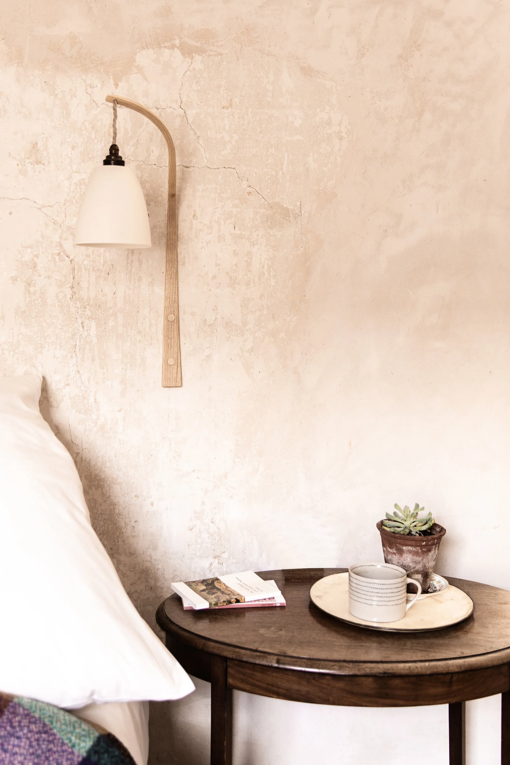 A round dark wooden side table holding three books, a white mug with black horizontal lines, and a tray with a potted succulent plant and a small cup, placed against a textured beige wall beside a bed with white bedding and a blanket with purple and 