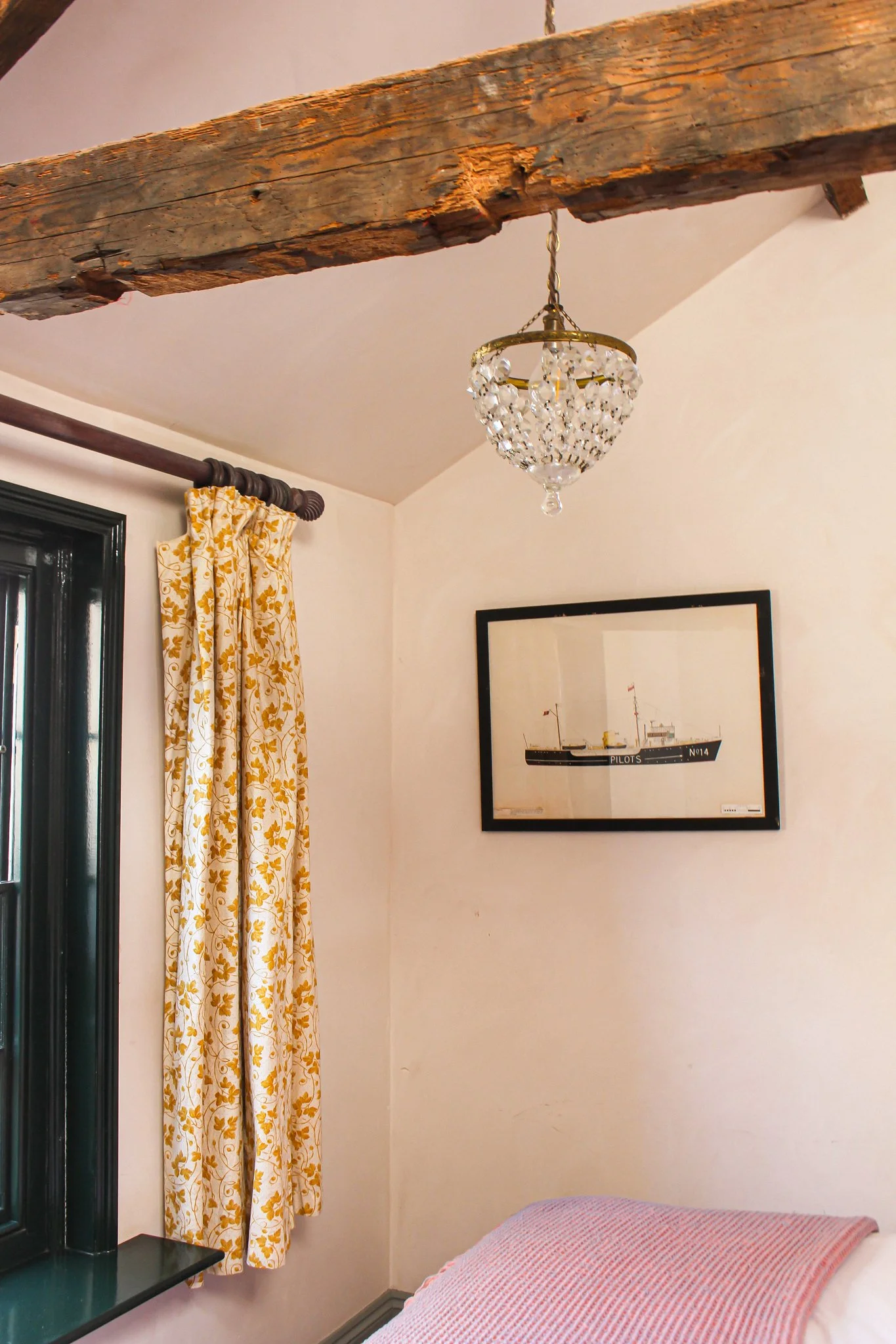 A bedroom corner with a wooden beam ceiling, a chandelier hanging from the ceiling, a window with yellow floral curtains, part of a bed with a pink and white striped bedspread, and a framed ship illustration on the wall.
