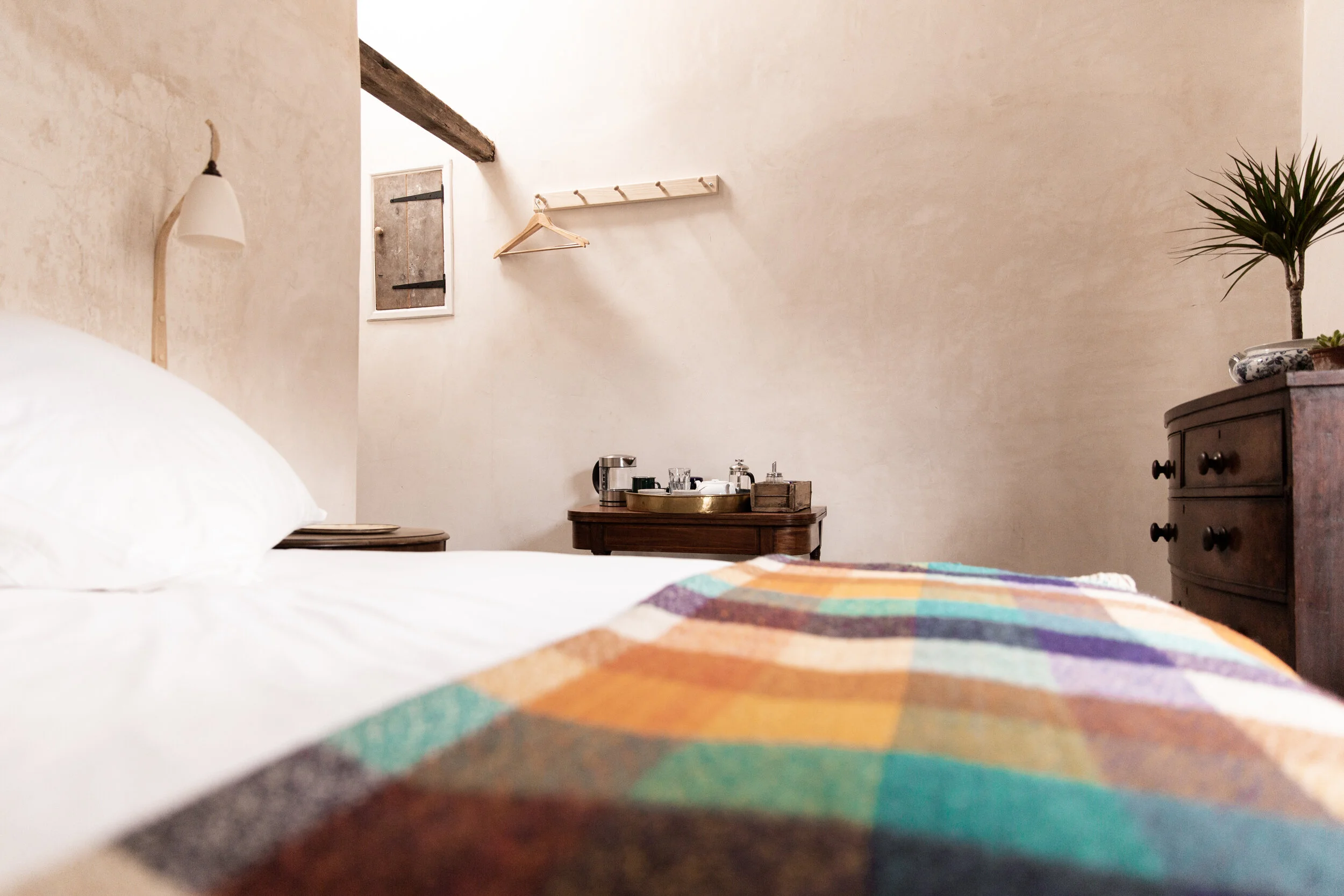 A cozy room with a bed covered in a colorful, striped quilt, a wooden side table with a tray holding a coffee maker and glasses, a dresser with a potted plant, a wooden coat rack with hangers, and a small window with rustic shutters.