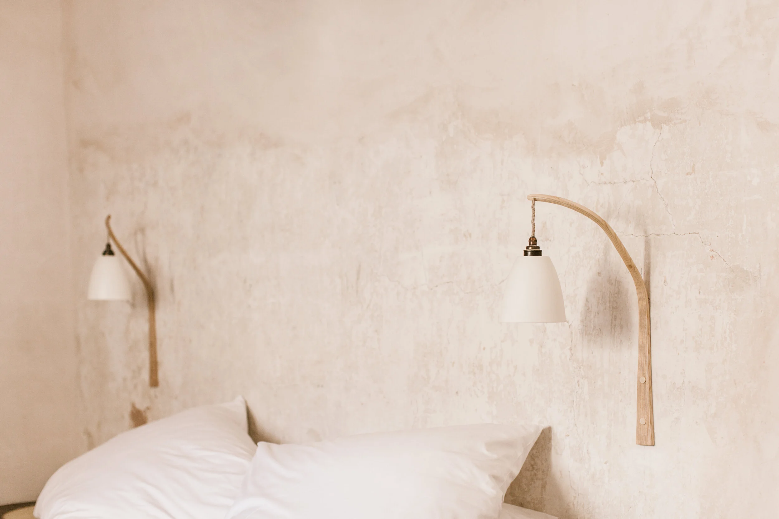 Two wall-mounted lamps with wooden arms and white shades above a bed with white pillows and bedding, against a textured, light-colored wall.
