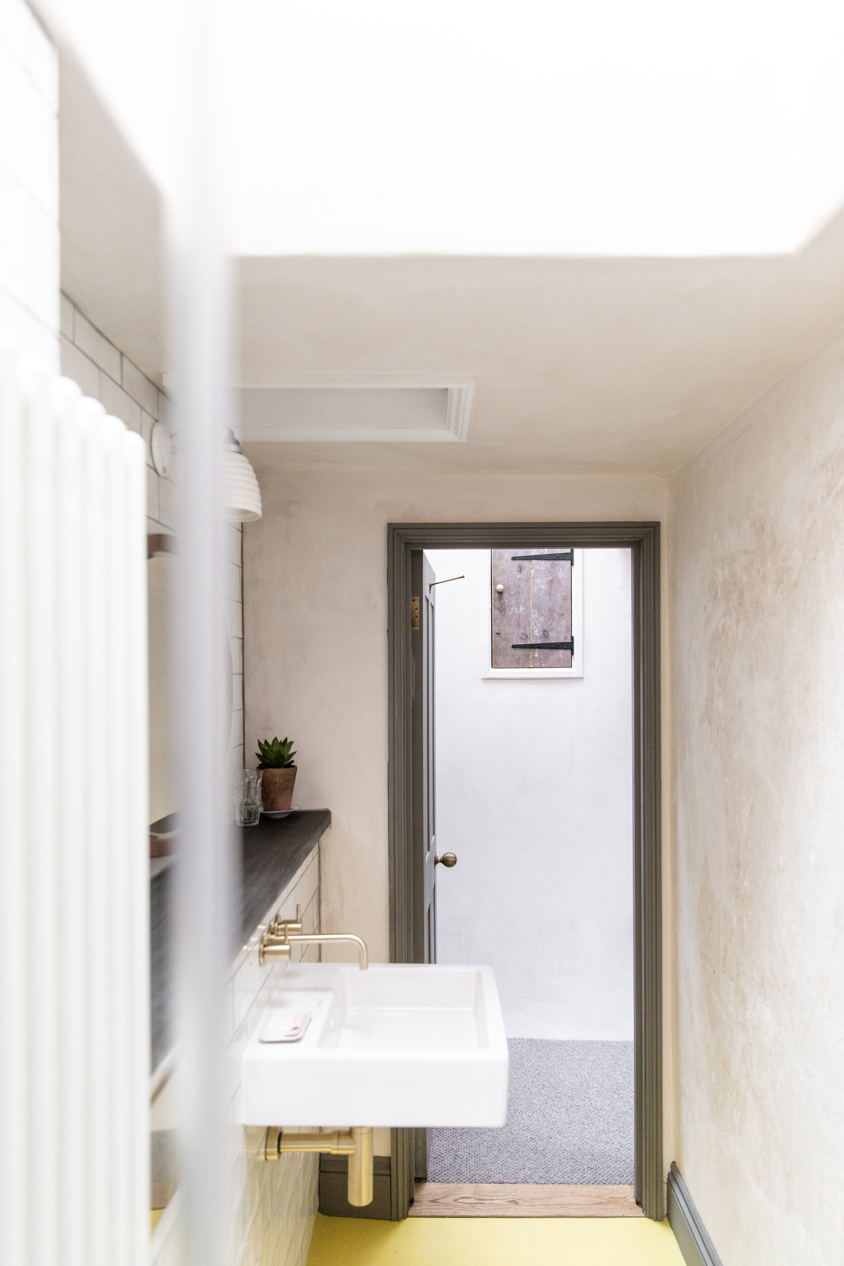 A small bathroom with a white sink, a small potted plant on the dark countertop, a gray door frame leading outside, a small window with a brown panel, and beige textured walls.