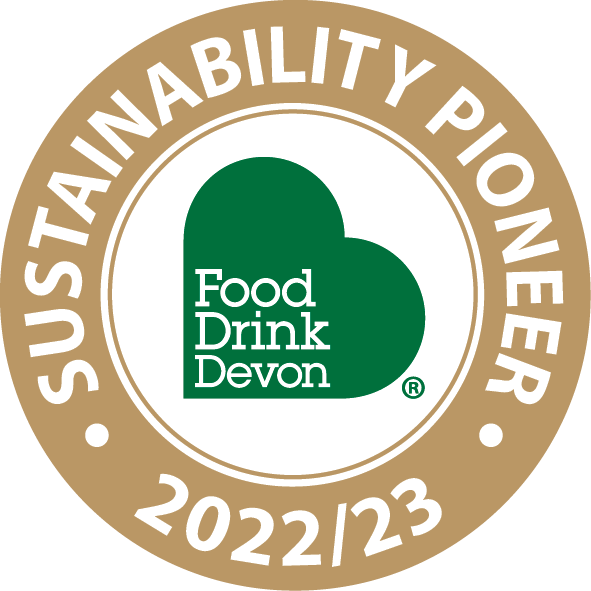 A circular badge with a beige outer ring and white inner circle. The outer ring has white text reading 'SUSTAINABILITY PIONEER 2022/23'. The center features a green heart shape with white text inside that says 'Food Drink Devon'.
