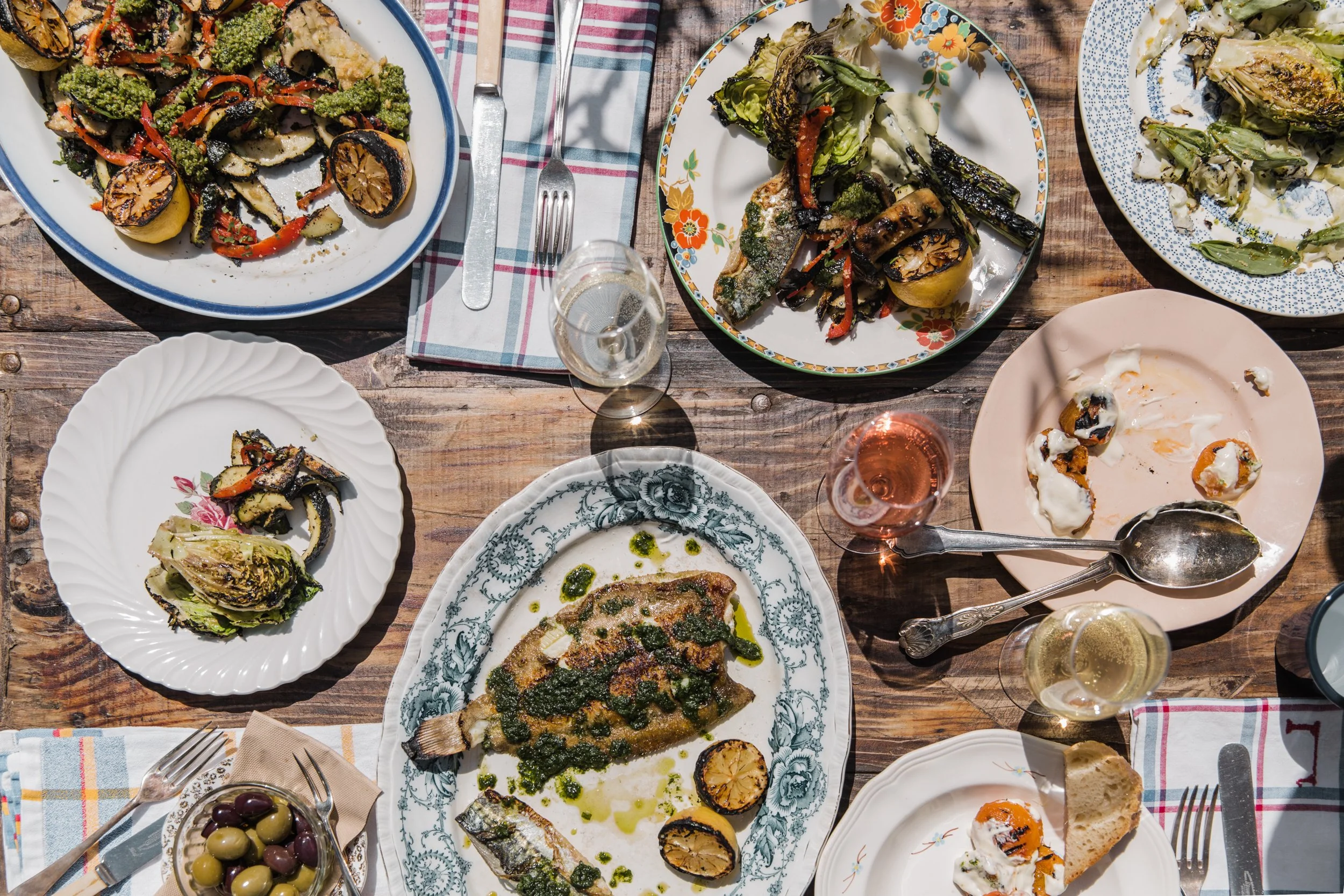 A variety of dishes on a wooden table, including grilled fish with herbs and lemon, roasted vegetables, a plate of sliced eggplant, and a bowl of green and black olives, alongside glasses of rosé wine and cutlery.
