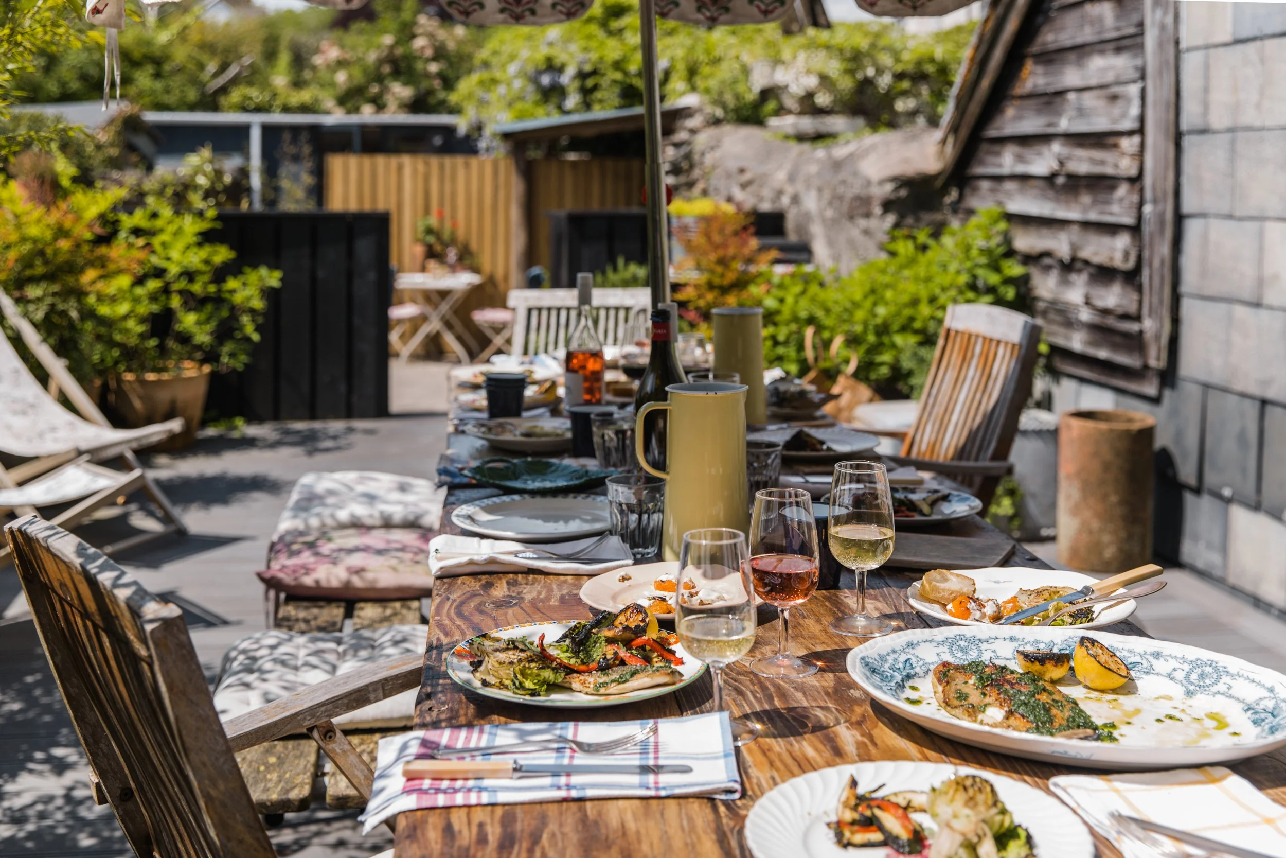 An outdoor dining table set with plates of food, glasses of wine, and mugs, surrounded by chairs and greenery, in a backyard setting.