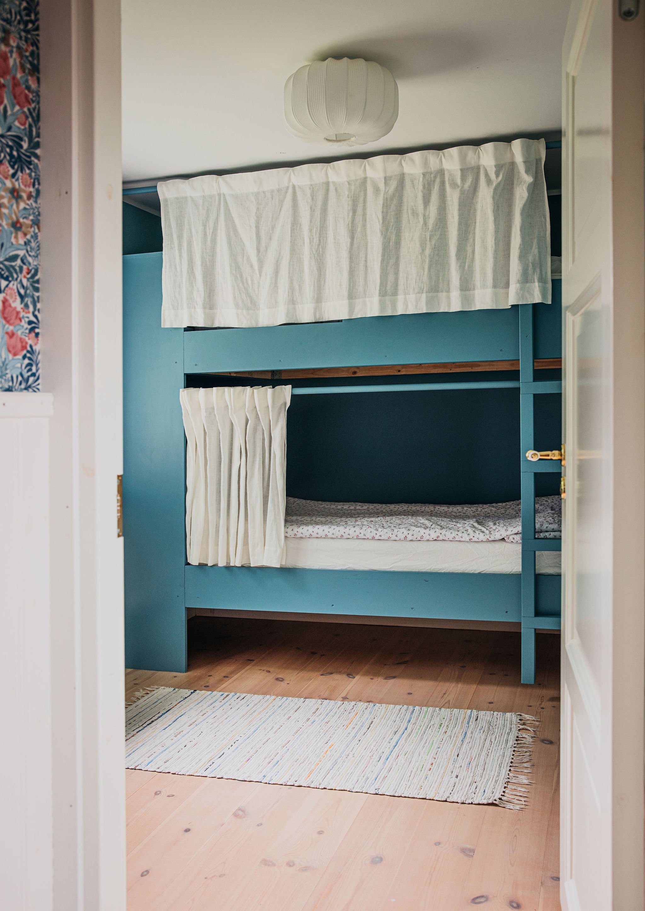 A view of two bunk beds in a small room, painted in blue, with white curtains covering the lower bunk and a white fabric hanging on top, a white ceiling with a round paper lantern light fixture, and a wooden floor with a colorful striped rug.