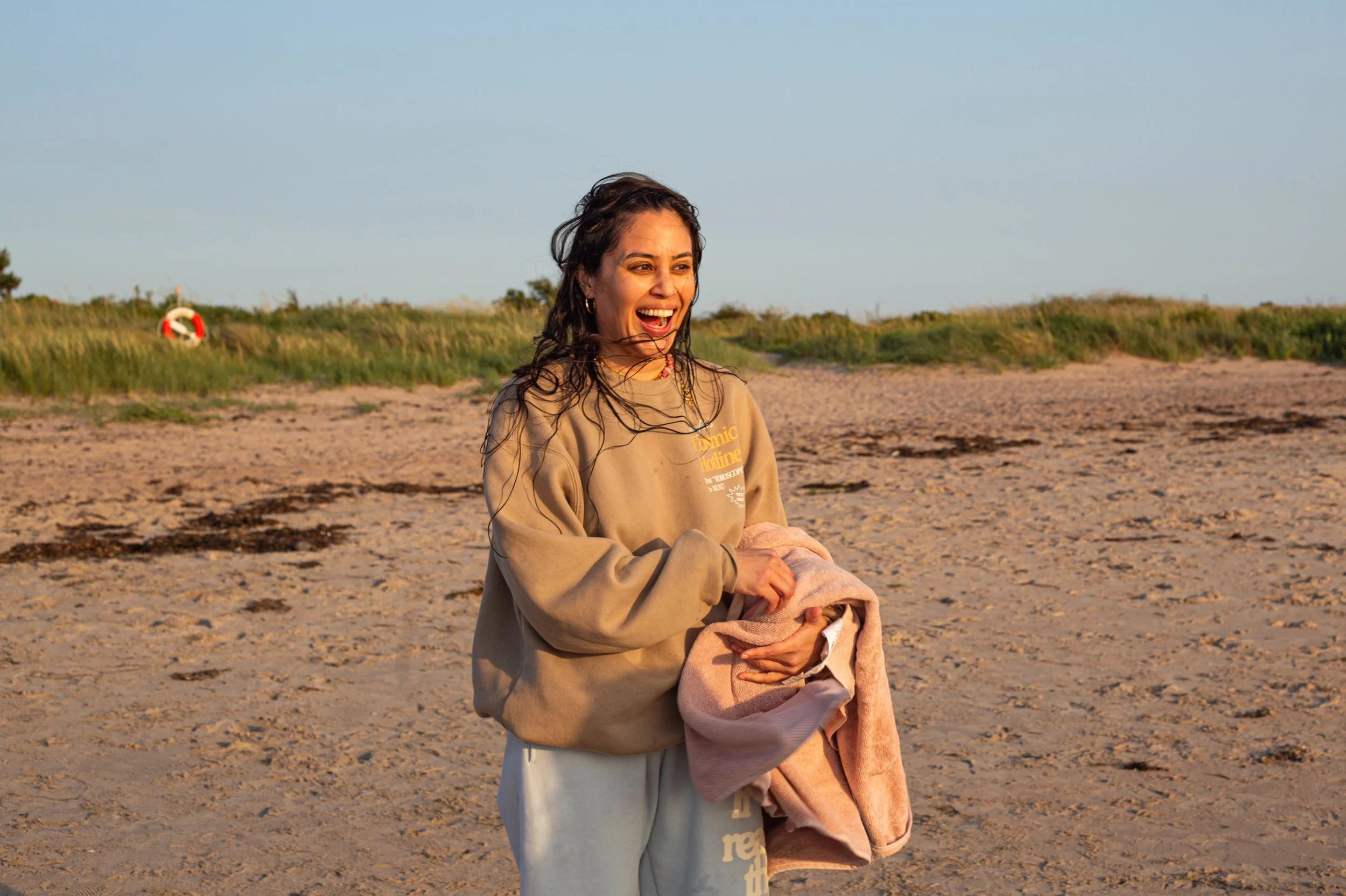 A young woman with shoulder-length dark hair, smiling and laughing on a sandy beach during sunset, holding a towel or blanket.