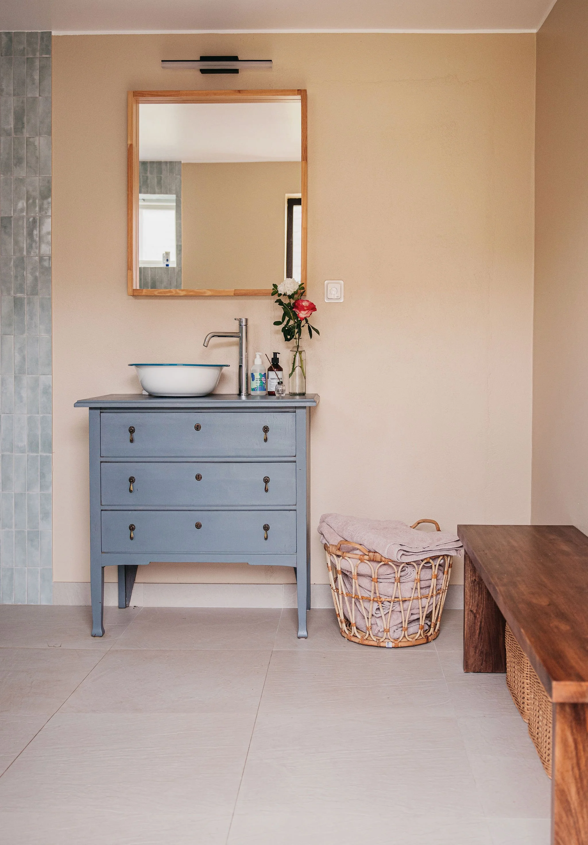 A blue dresser with a vessel sink, a tall faucet, a mirror, a soap dispenser, a vase with flowers, a basket with folded towels, and a wooden table in a bathroom with beige and gray walls and beige tile flooring.