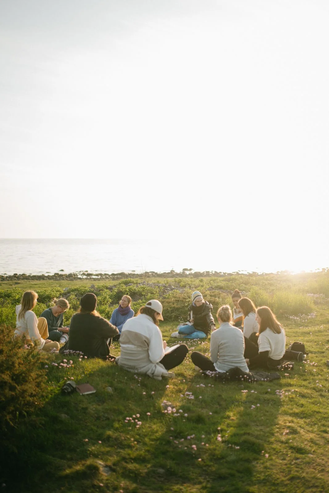 A group of people sitting in a circle on grass outdoors near the water, enjoying the sunset.
