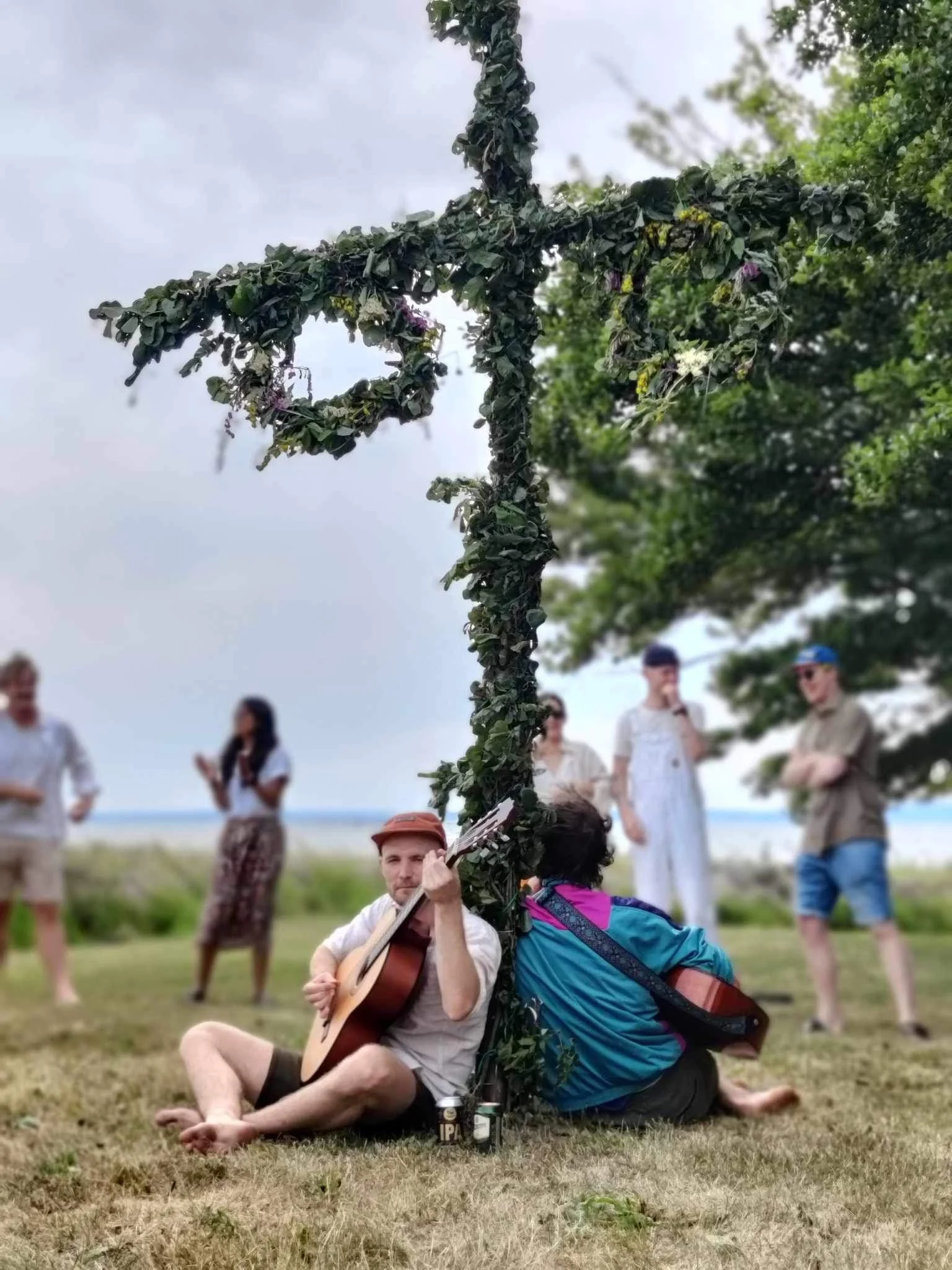 A man with a guitar sitting on the ground, surrounded by a group of people outdoors near a beach. A large, decorative cross made of greenery is in front of him. The background shows trees and a cloudy sky.