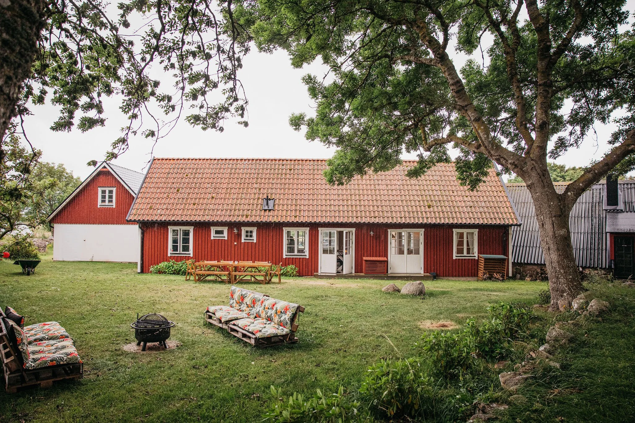A red house with a sloped roof, surrounded by green grass and trees. There are outdoor chairs and a table in the yard, and a large tree in the foreground with branches extending over the yard.