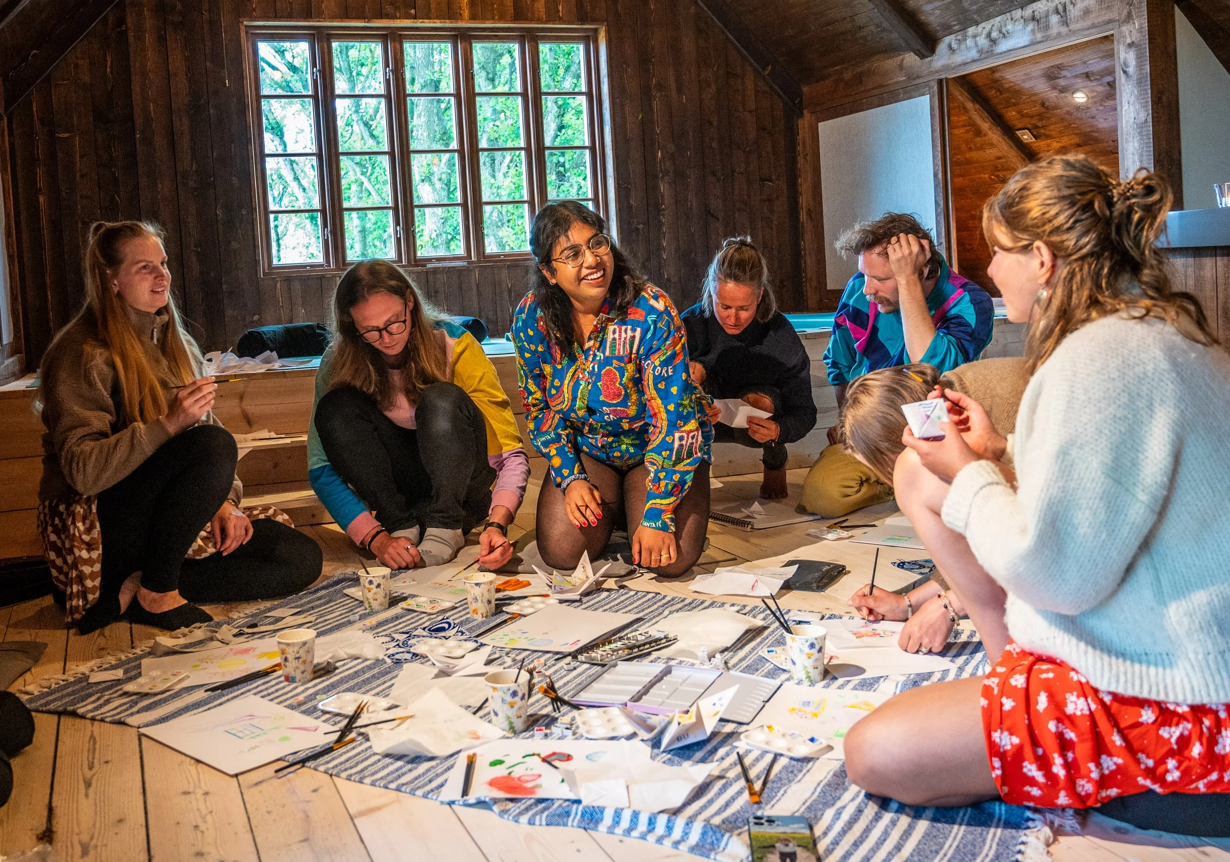 Group of women sitting on the floor, engaging in arts and crafts, with painting supplies and paper scattered on a blue and white striped cloth.