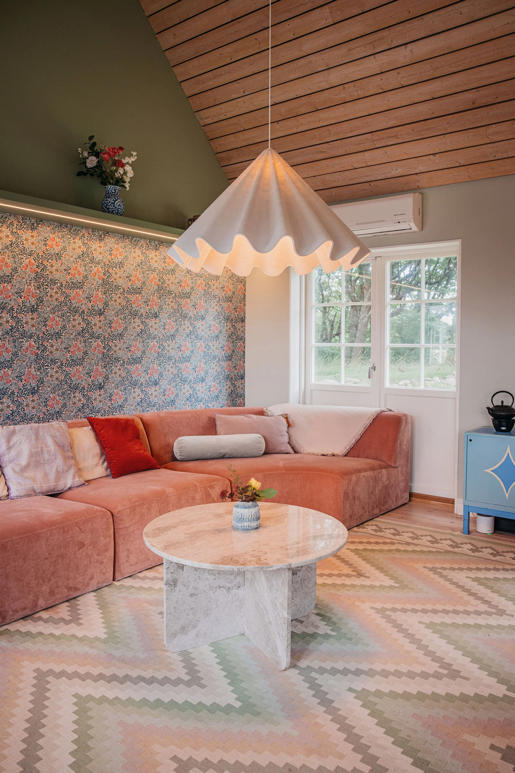 Living room with a pink velvet sectional sofa, a marble coffee table, a patterned rug, a white pleated pendant lamp, large windows, a floral wallpaper accent wall, a blue cabinet, and a wood-paneled ceiling.
