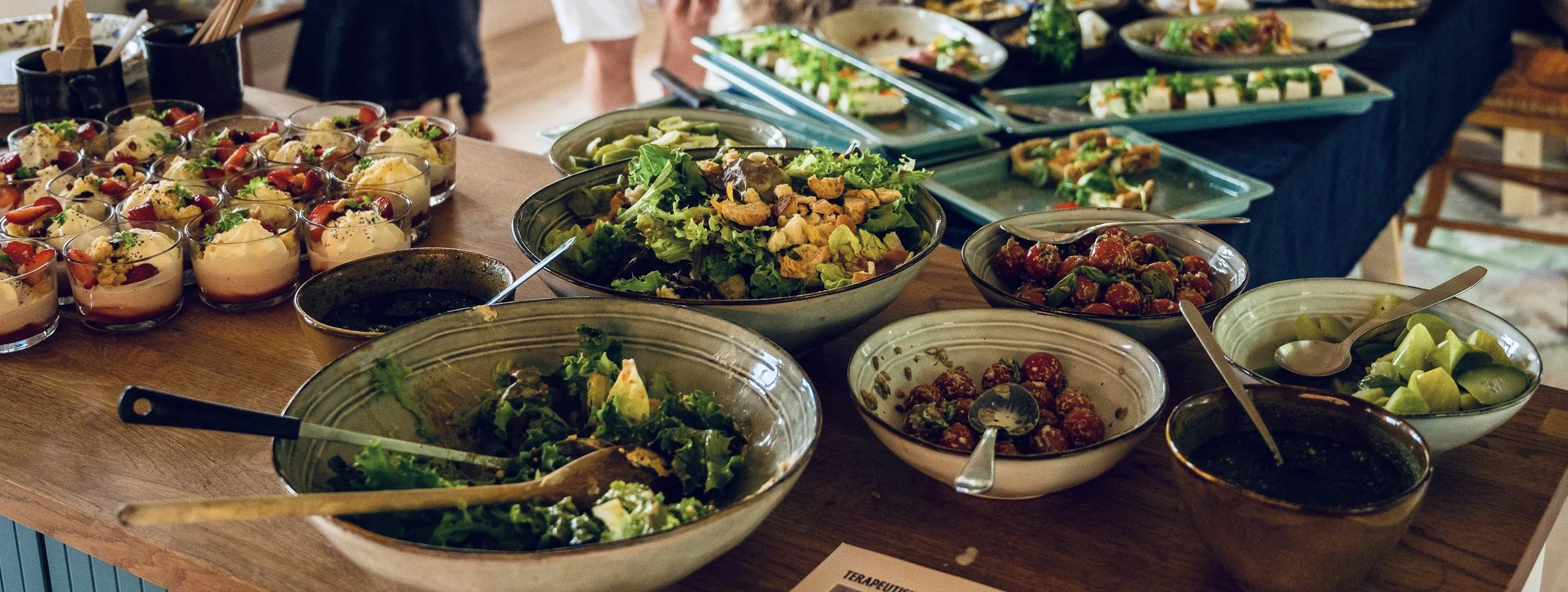 A variety of salads and appetizers on a wooden table, including stuffed strawberries, green beans, mixed greens, cherry tomatoes, and other vegetable dishes, with small serving utensils in some bowls.
