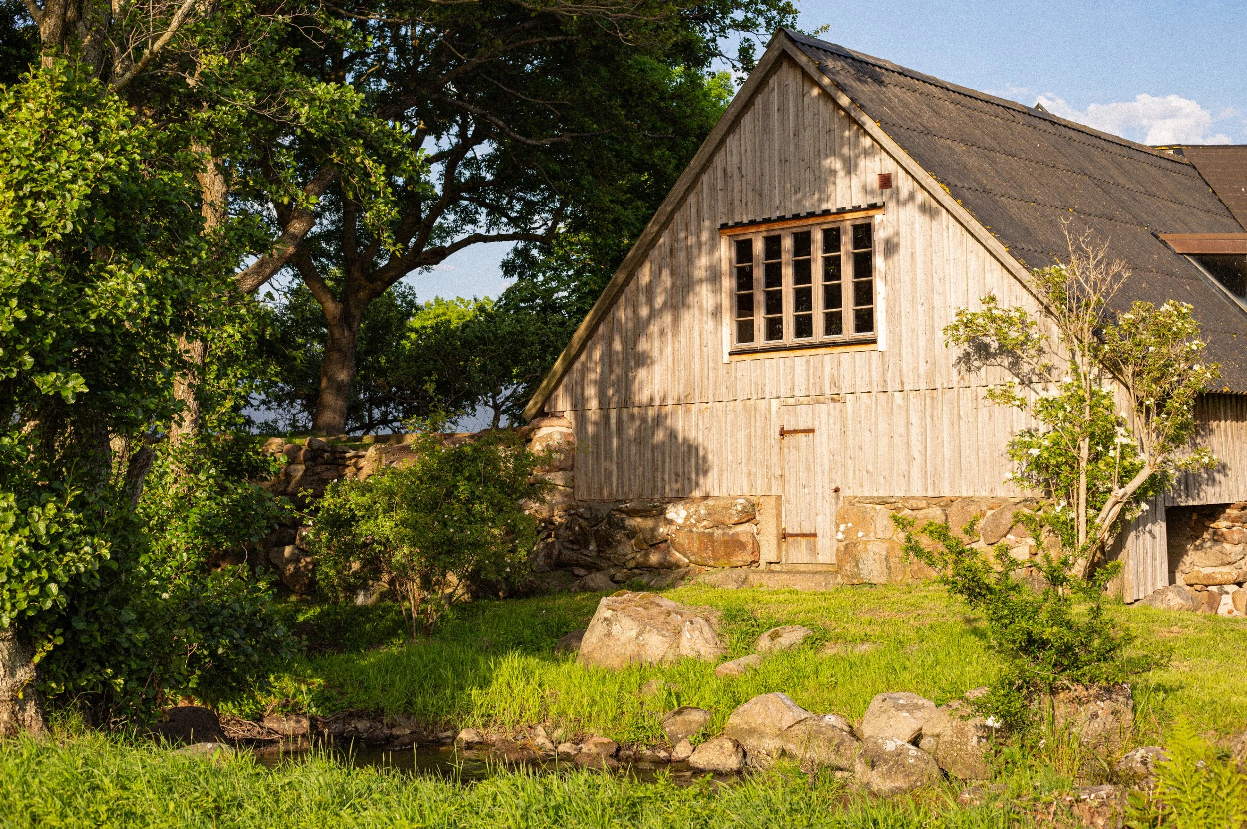 A rustic wooden barn with a sloped roof next to a large leafy tree, surrounded by rocks and green grass, under a blue sky with some clouds.