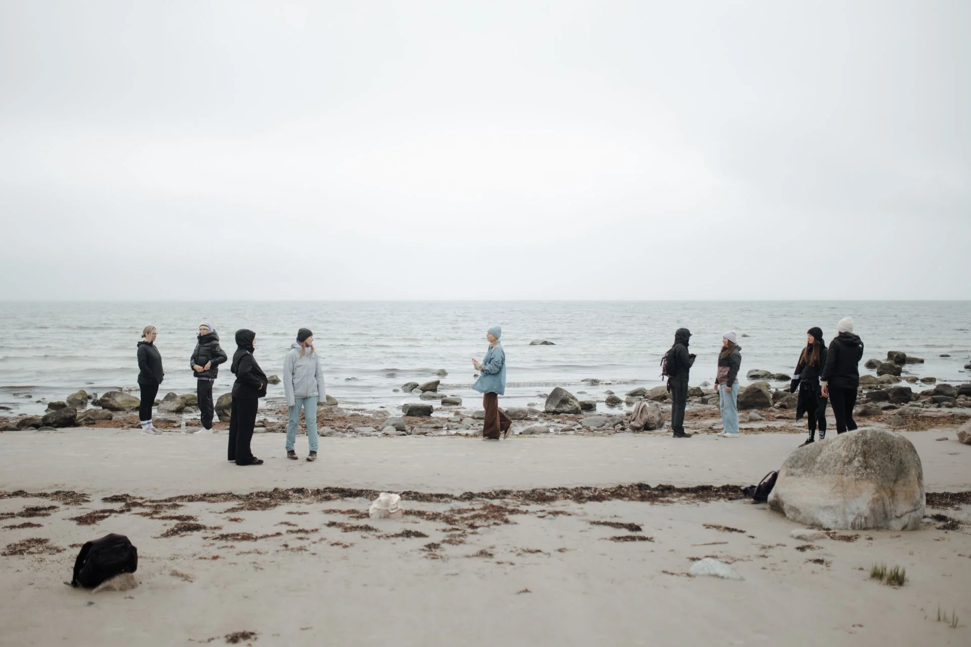 A group of people standing on a beach, facing the ocean on a cloudy day, with rocks scattered along the shore, dressed in jackets and warm clothing.