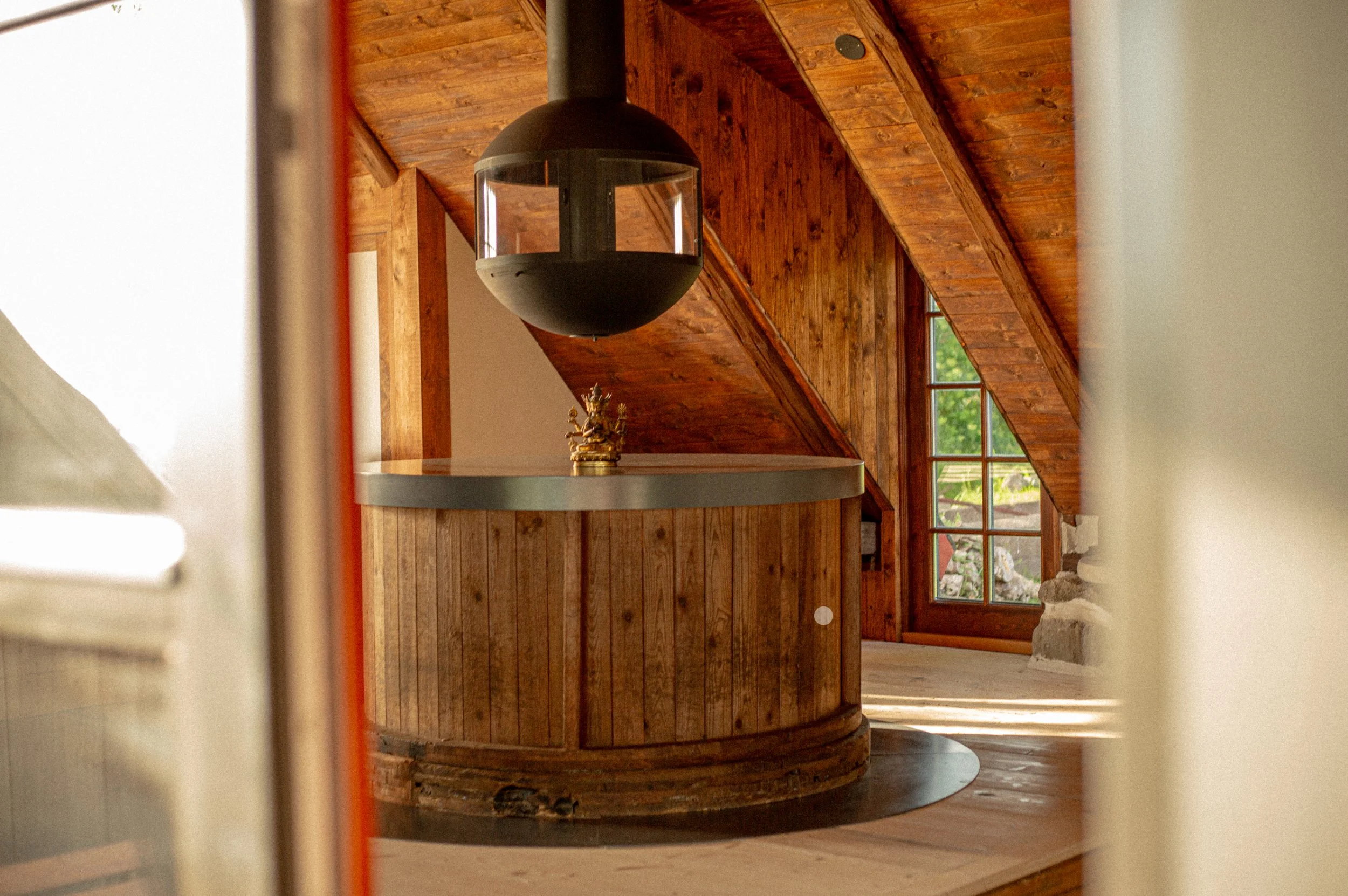 Interior view of a wooden room with large windows and a round wooden bar. A black spherical hanging light fixture is in the center of the image.