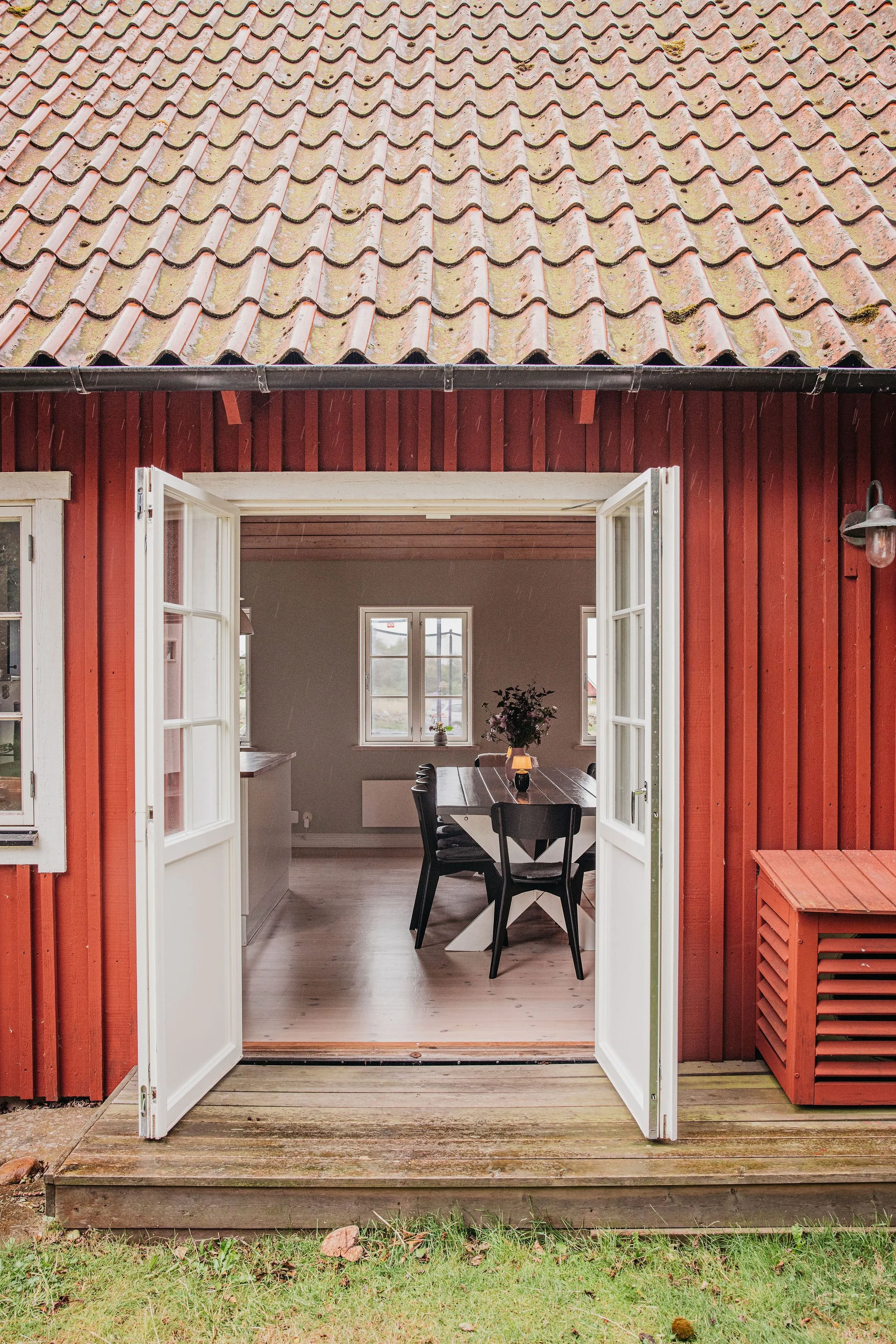 View of a house with red wooden exterior walls and a sloped, tiled roof. French doors are open, revealing a dining area inside with a wooden table, black chairs, and a window with natural light. There is a small deck outside with grass and pebbles on
