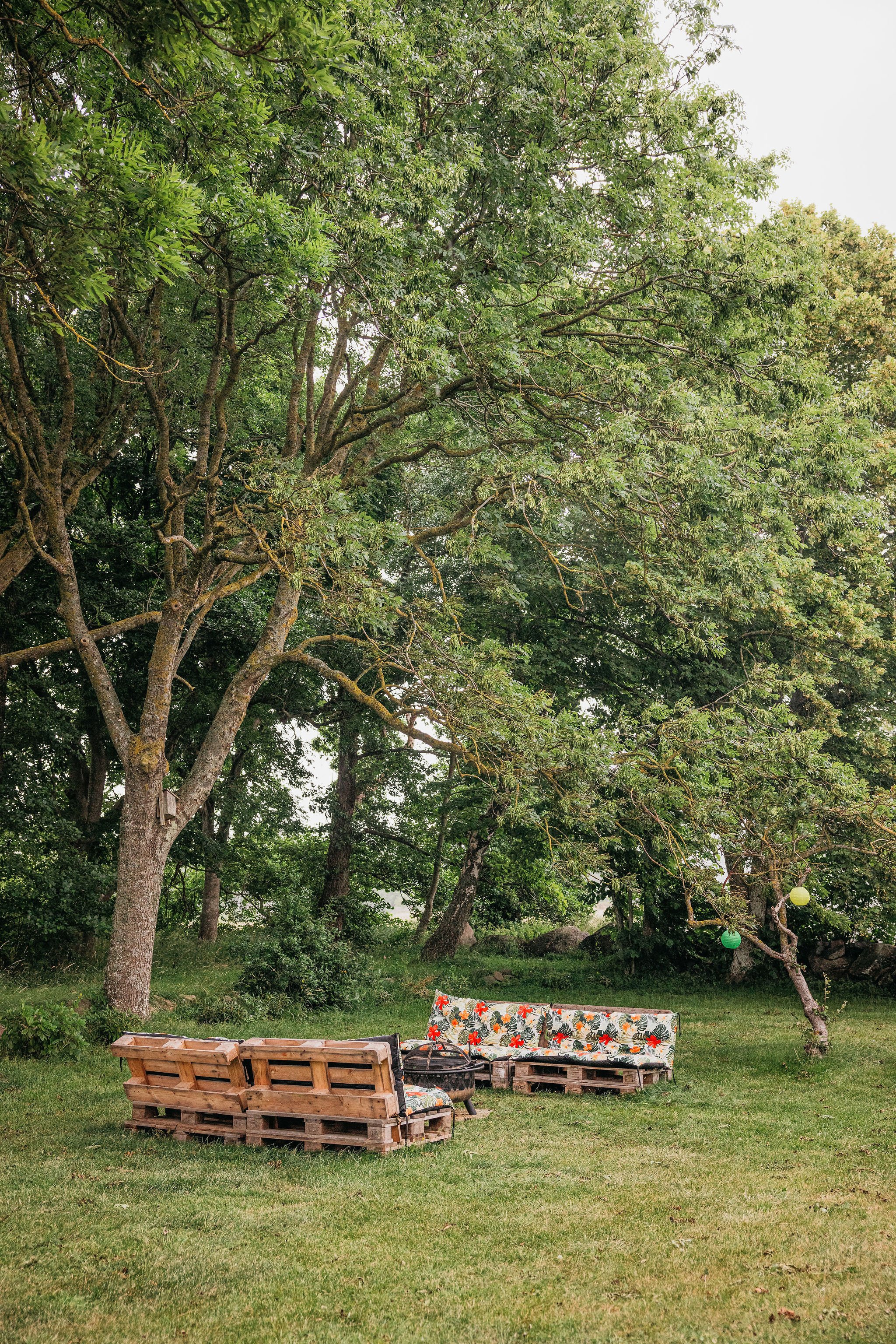 A backyard outdoor scene showing a wooden pallet sofa with floral cushions under trees, string lights hanging between trees, and a grassy area.