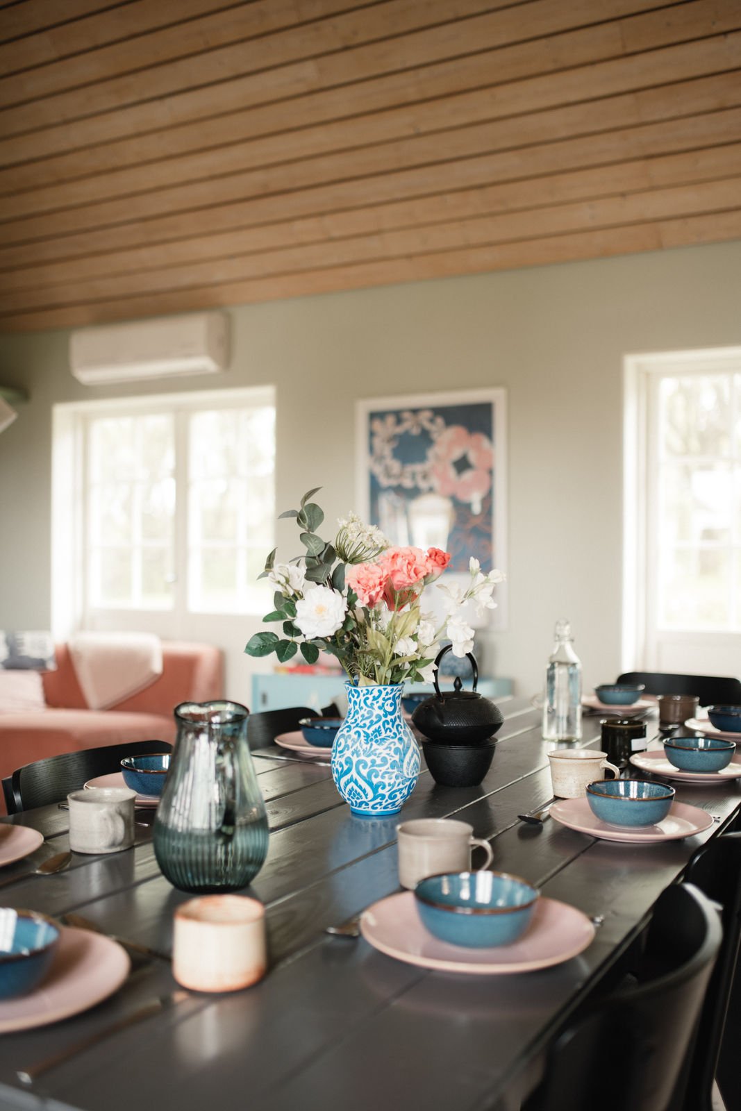 A dining table set with pink and blue dishes, bowls, and mugs, featuring a blue and white patterned vase with pink and white flowers, in a bright room with windows and wall decor.