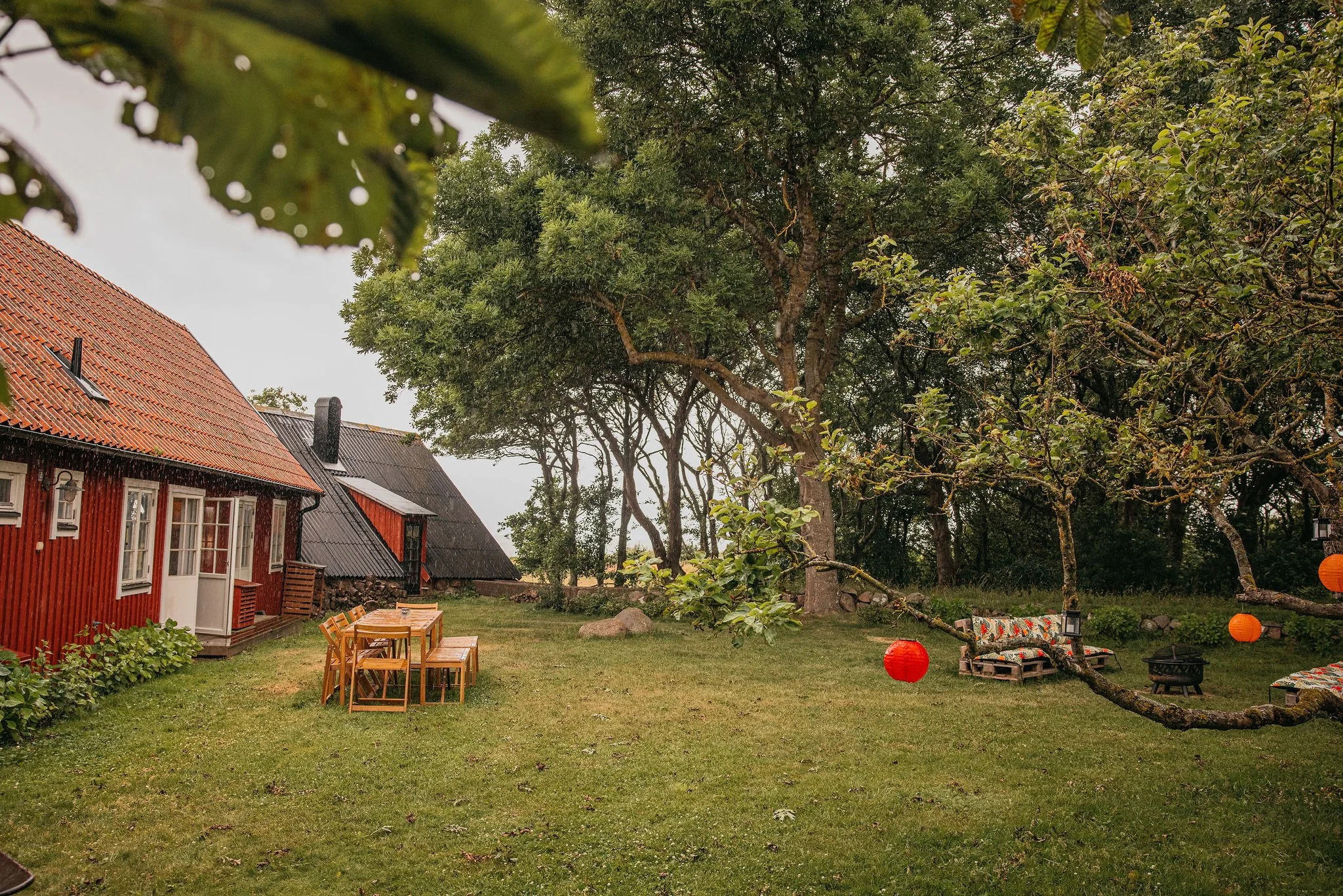 A backyard scene with a red wooden house, trees, a green lawn, outdoor furniture, orange lanterns, and a large tree.
