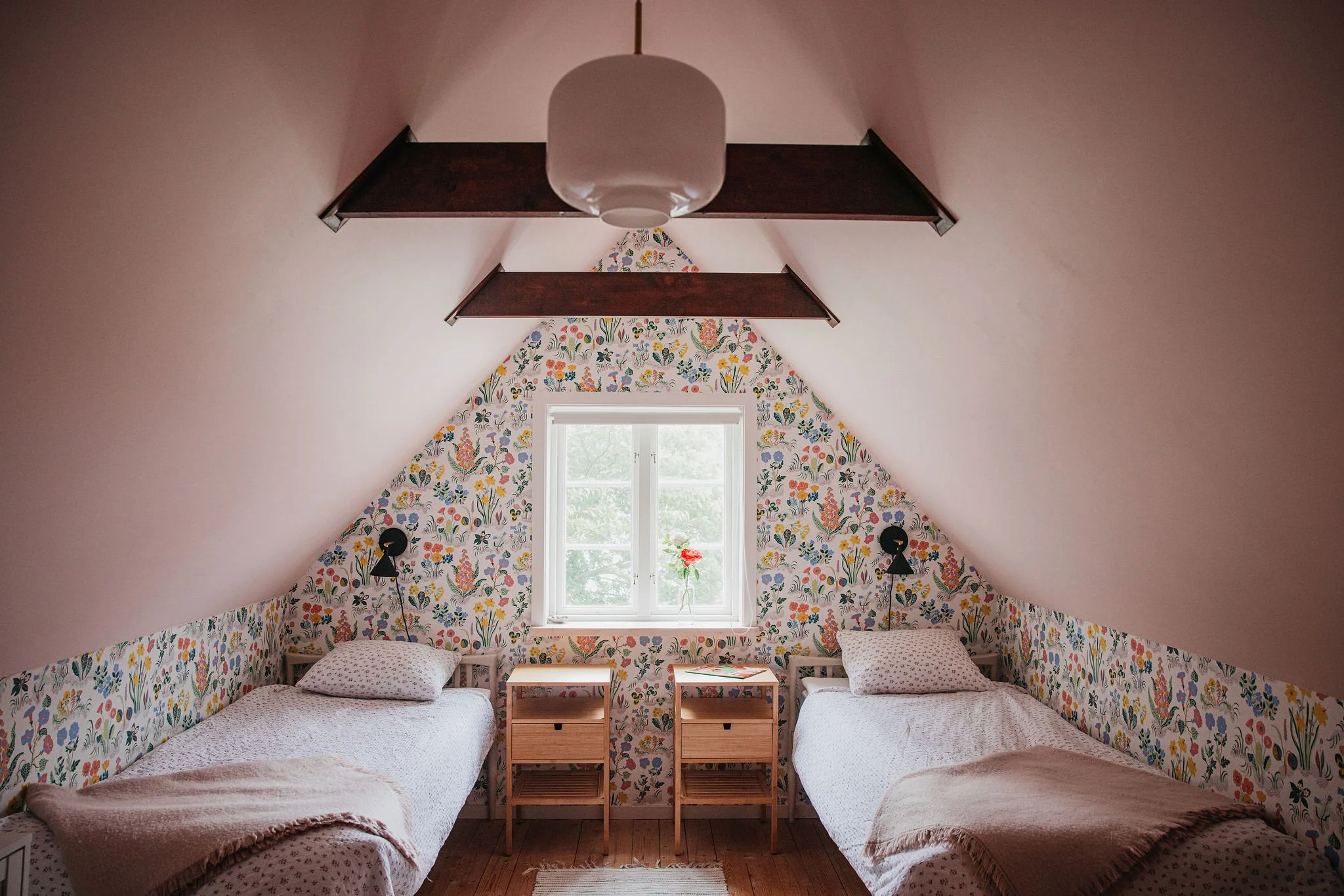 A cozy attic bedroom with two beds, floral wallpaper on the back wall and a window in the center, sunlight coming through, and small nightstands between the beds.