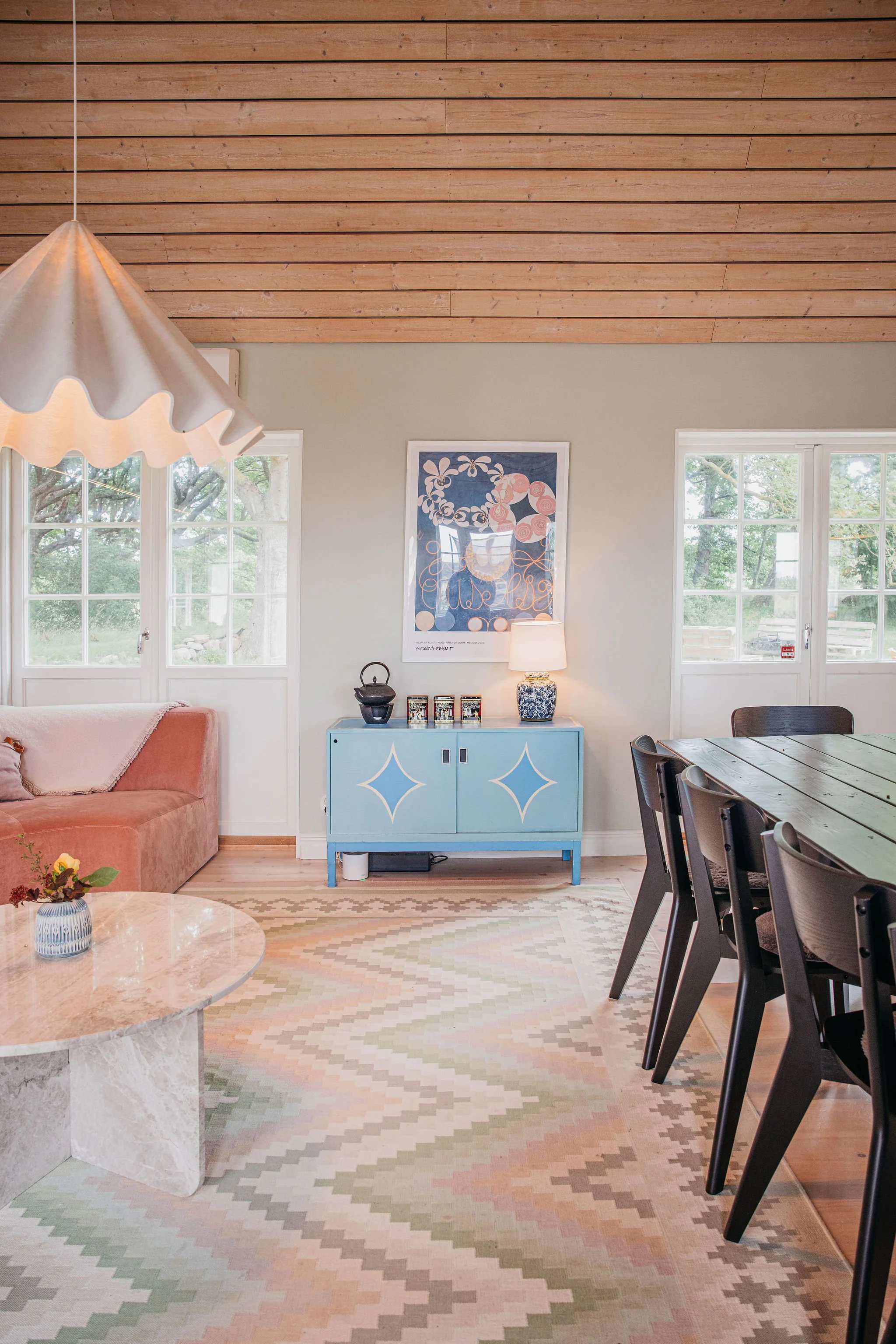 Interior of a living room with a wooden ceiling, a pink sofa, a marble coffee table with a small vase of flowers, a light blue cabinet with decorative items, artwork on the wall, and large windows letting in natural light.