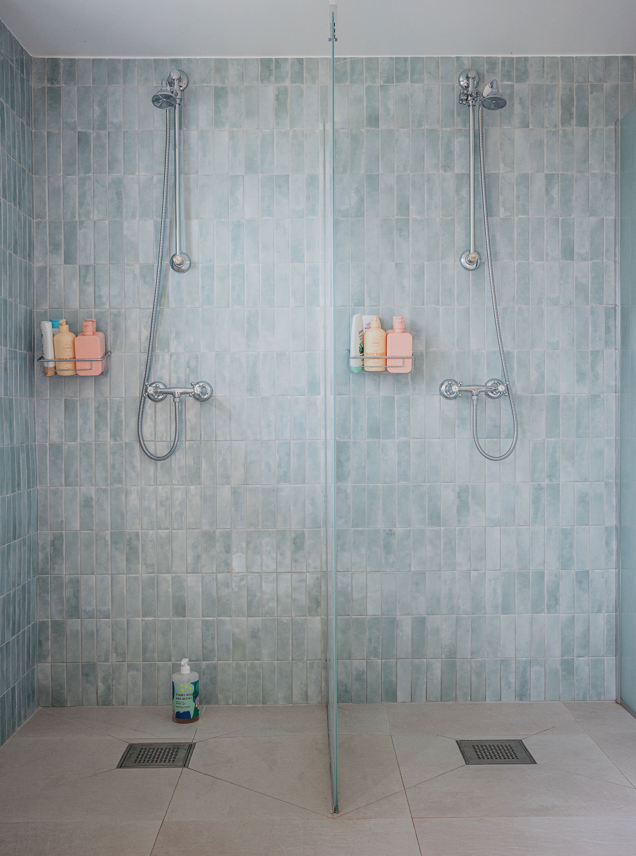 A modern communal shower with two showerheads, a clear glass divider in the center, and a tiled wall. There are shelf racks with bottles of shampoo and body wash. A large container of body lotion is on the floor in front of the left drain.