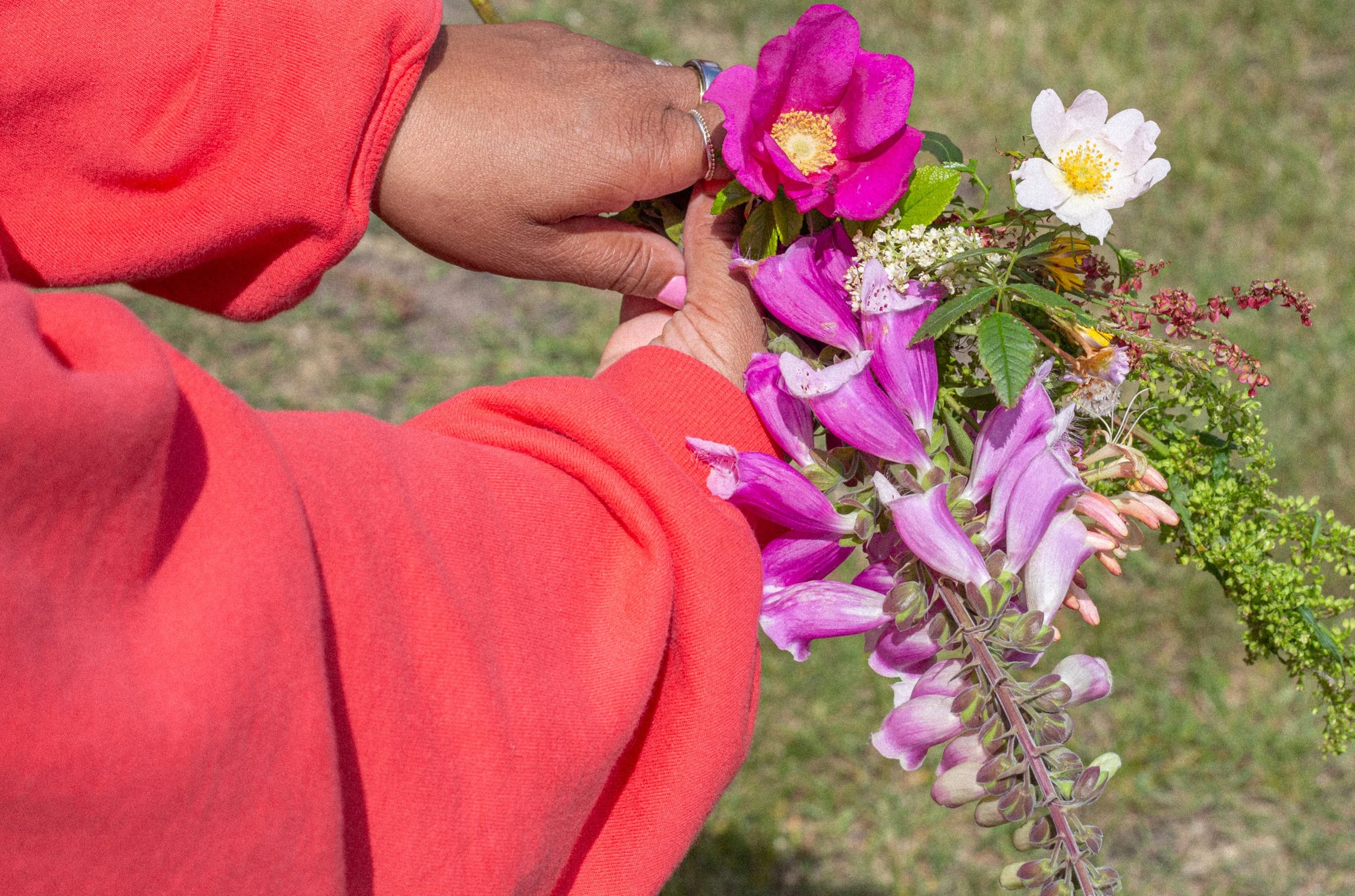 Person holding a bouquet of colorful flowers, including pink, white, and purple blossoms, wearing an orange sweater and silver jewelry, with a grassy background.