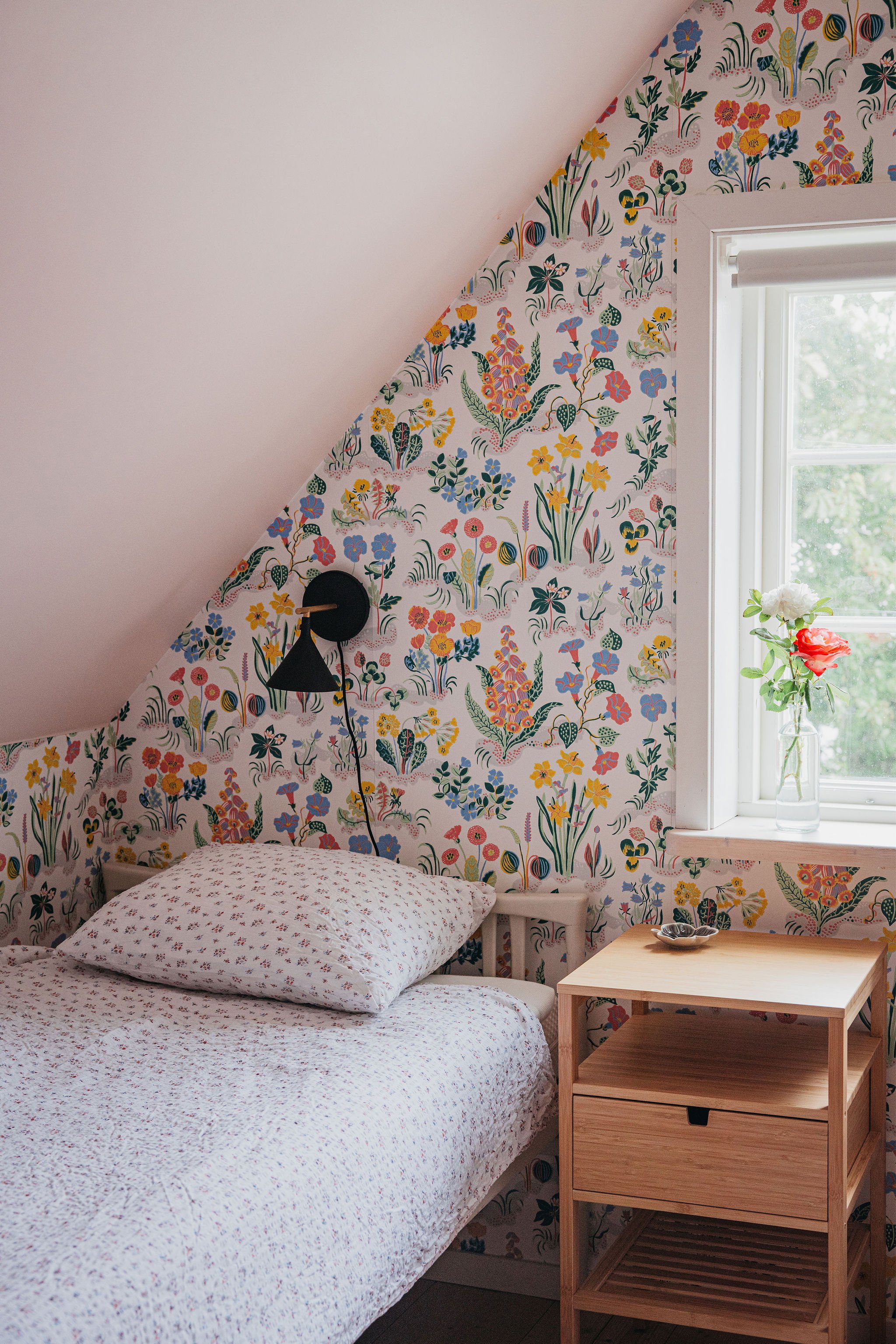 A cozy bedroom with floral patterned wallpaper on an angled wall, a white bed with matching floral bedding, a black wall-mounted reading lamp, a wooden nightstand with open shelves, a window with a flower vase, and greenery visible outside.
