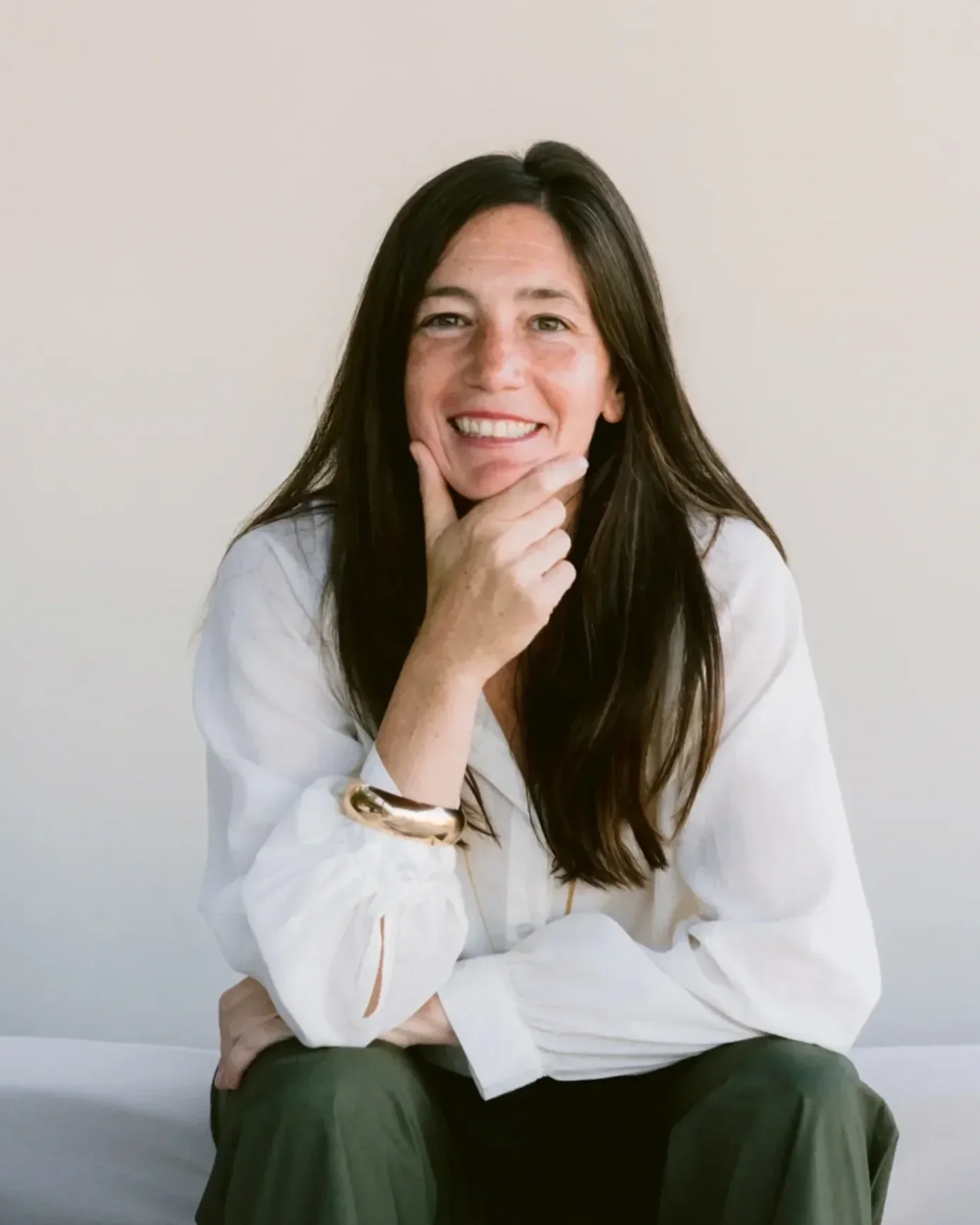 A woman with long dark hair smiling, wearing a white blouse with rolled-up sleeves, dark green pants, and a gold bracelet on her right wrist, sitting against a plain light-colored background.