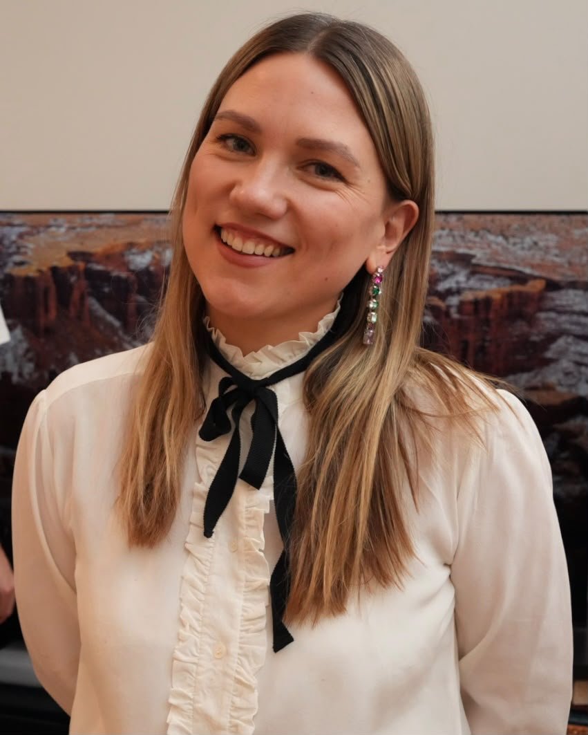 A woman with long blonde hair smiling, wearing a cream-colored blouse with ruffles and a black ribbon tied in a bow, and a colorful earring with green, pink, and purple stones.