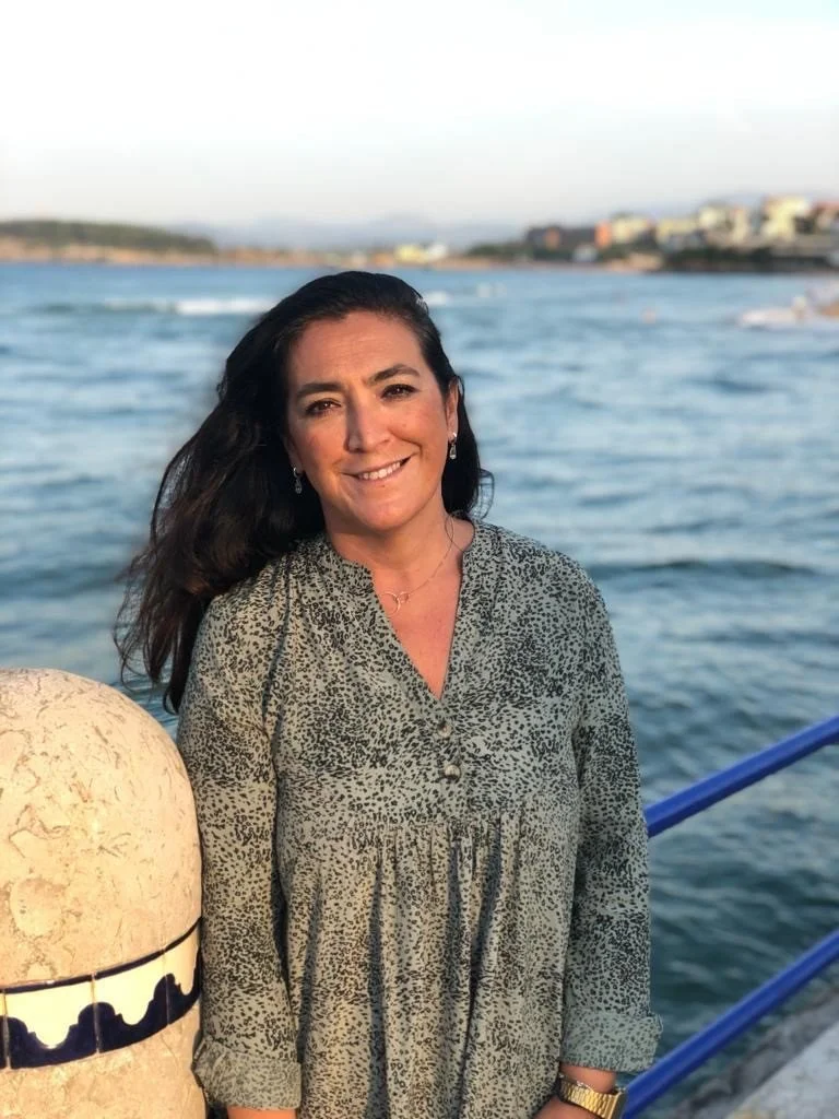 A woman with dark hair smiling near a body of water with buildings in the background.